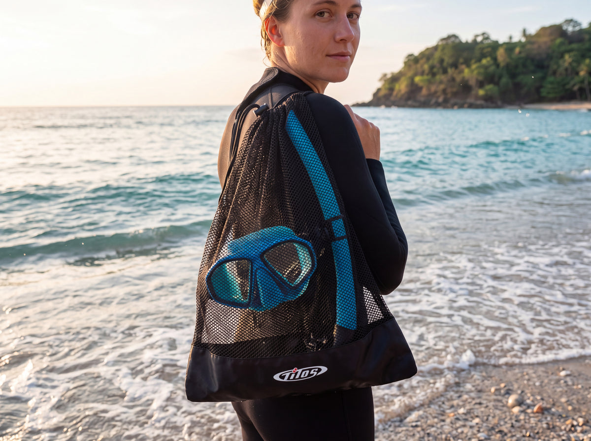 A snorkeler carries a black Tilos mesh mask and snorkel bag over one shoulder while standing in shallow surf at golden hour.