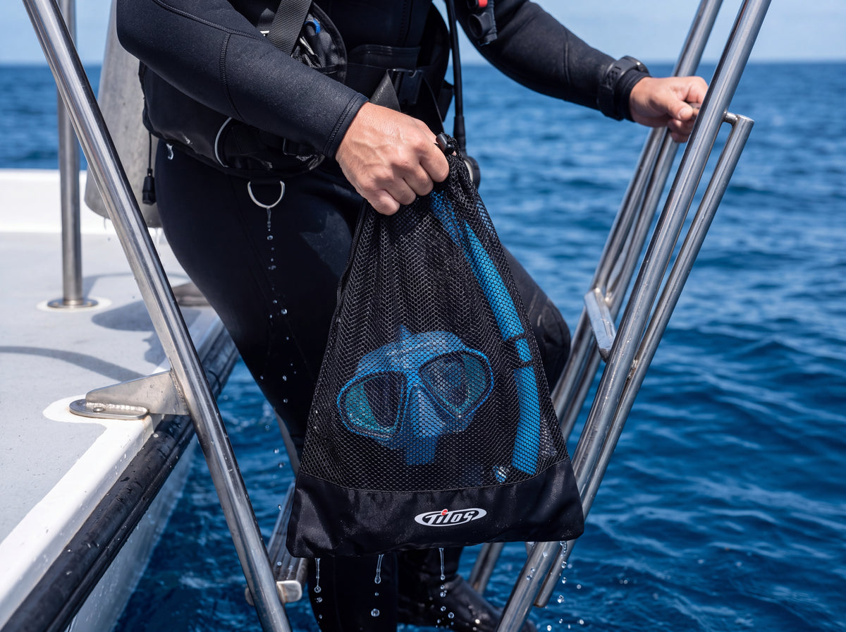 A diver climbs a stainless boat ladder holding a black Tilos mesh mask and snorkel bag draining seawater.