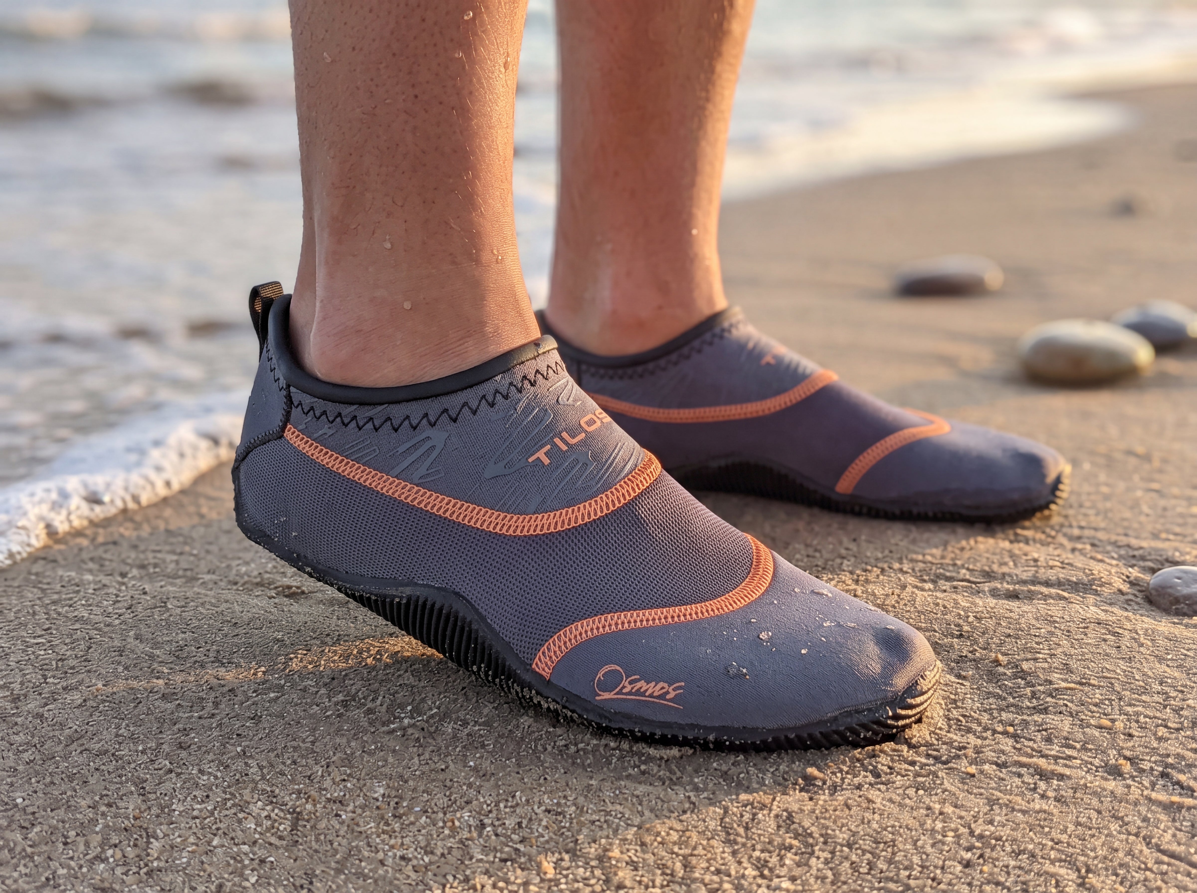 Gray Tilos Osmos water socks being worn at the shoreline in golden hour light, front three quarter view showing the TILOS wordmark on the ankle collar