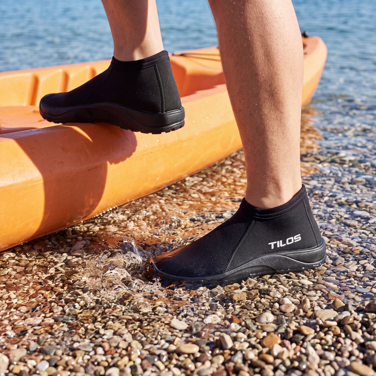 Kayaker stepping out of an orange kayak onto a pebbly shoreline while wearing the black Tilos 3mm Low Cut Molded Vulcanized Sole Beach Boot