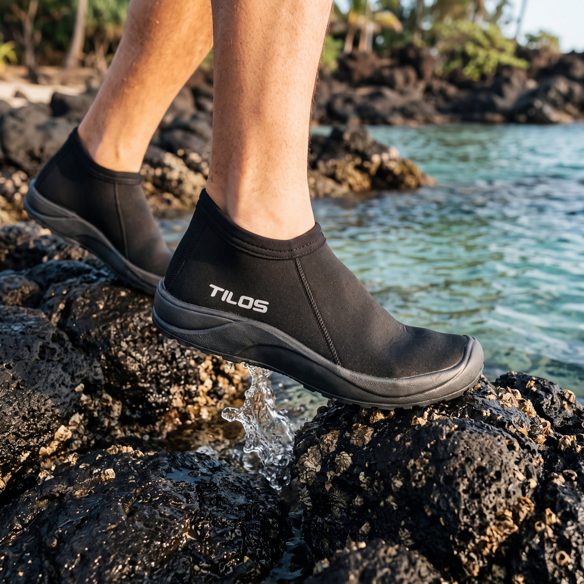 Person stepping across wet volcanic rocks by a tide pool while wearing the black Tilos 3mm Low Cut Molded Vulcanized Sole Beach Boot