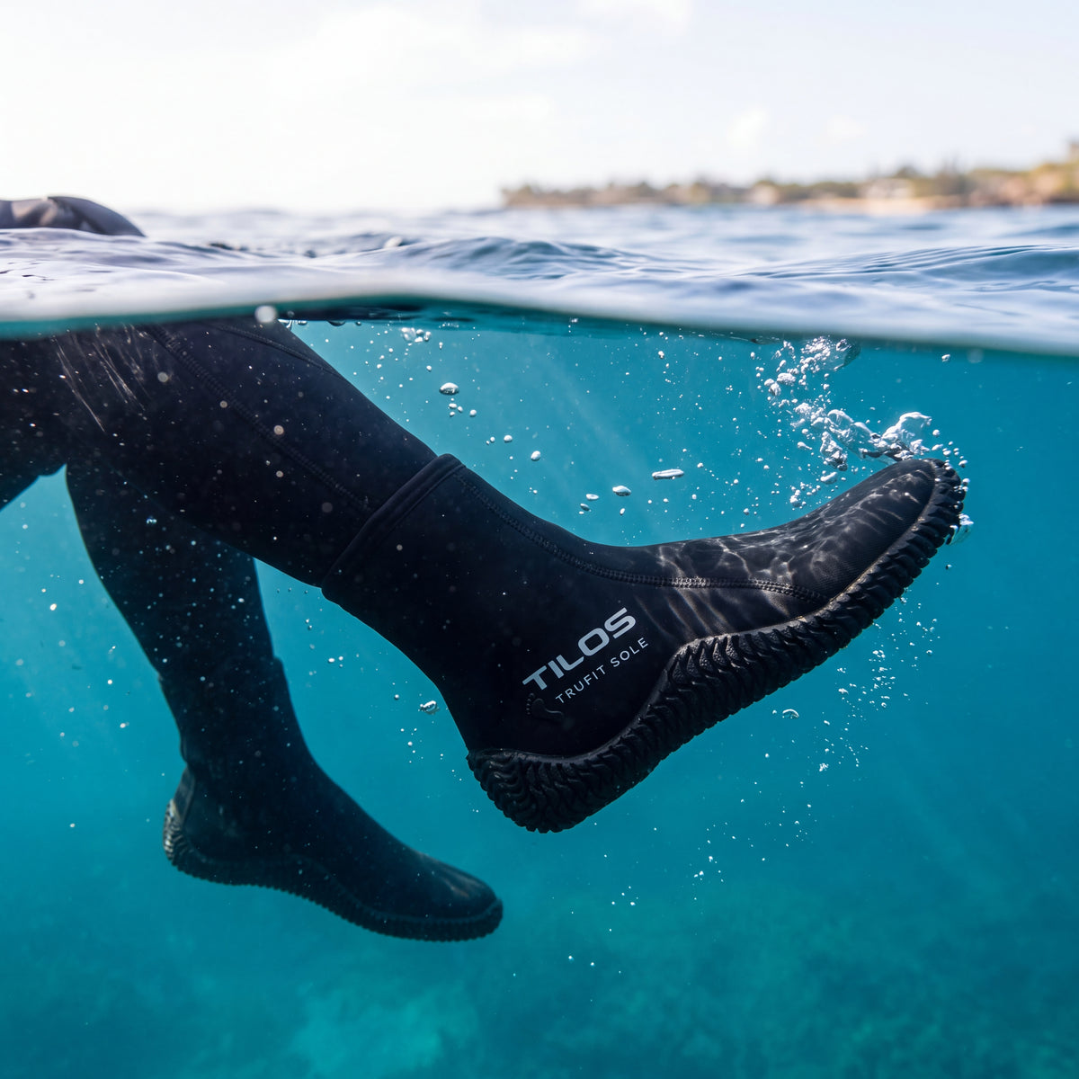 A snorkeler floating at the surface of clear tropical water, wearing Tilos 5mm Titanium Zip Trufit boots, photographed from the waterline with the silver Tilos ankle logo visible beneath the split.