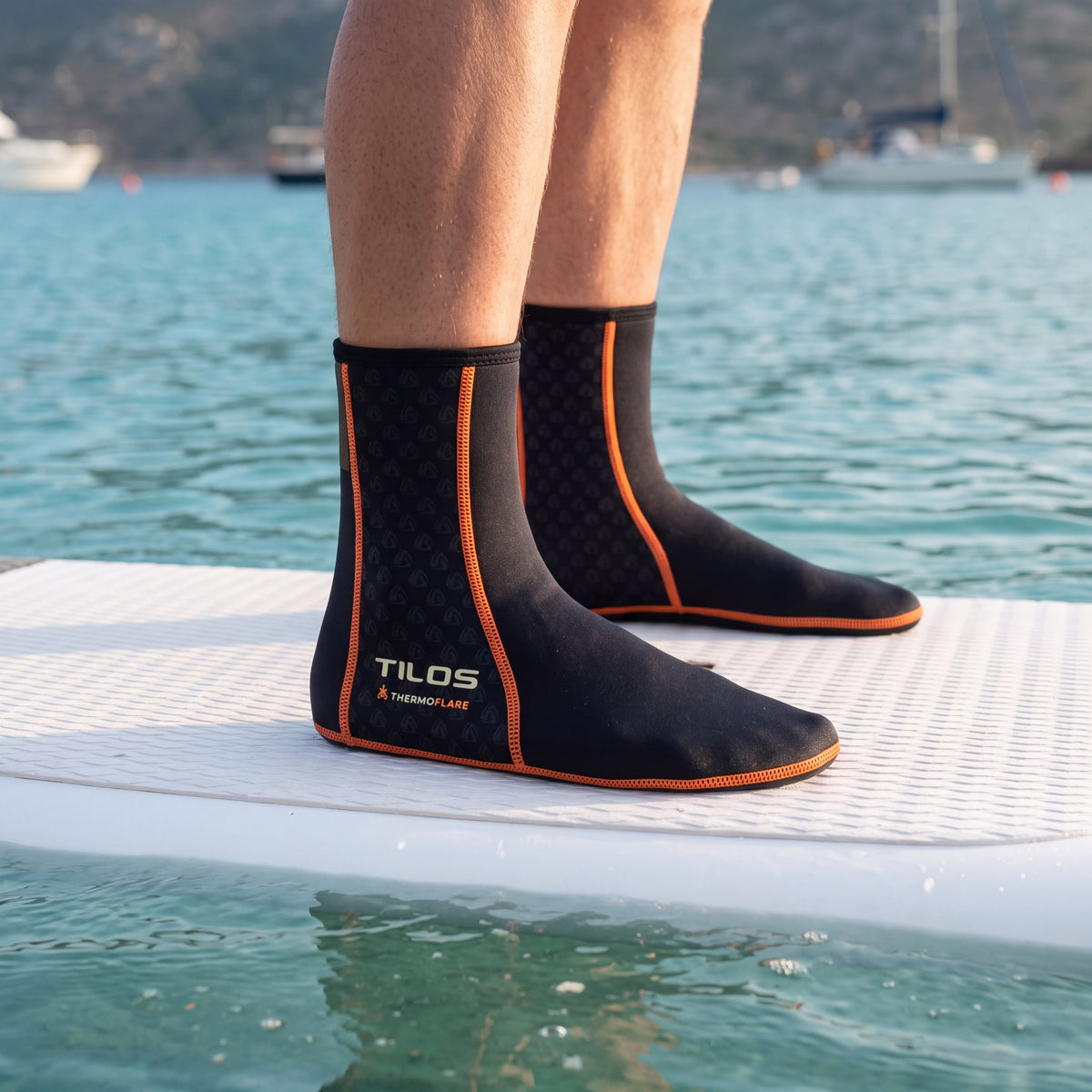 Paddleboarder standing on a white board in a calm bay while wearing the black Tilos 1mm Thermoflare Socks