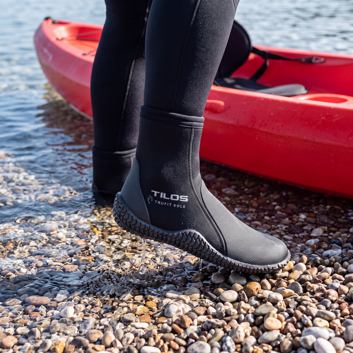 Kayaker stepping ashore beside a red kayak on a pebbly shoreline while wearing the black Tilos 5mm Thermoflare Semi-Dry Trufit Boot
