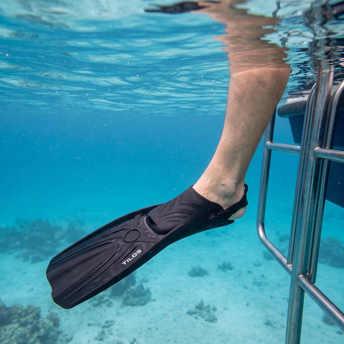 Diver at a boat ladder with one bare leg hanging into clear blue water, wearing a black Tilos Aubade open heel fin