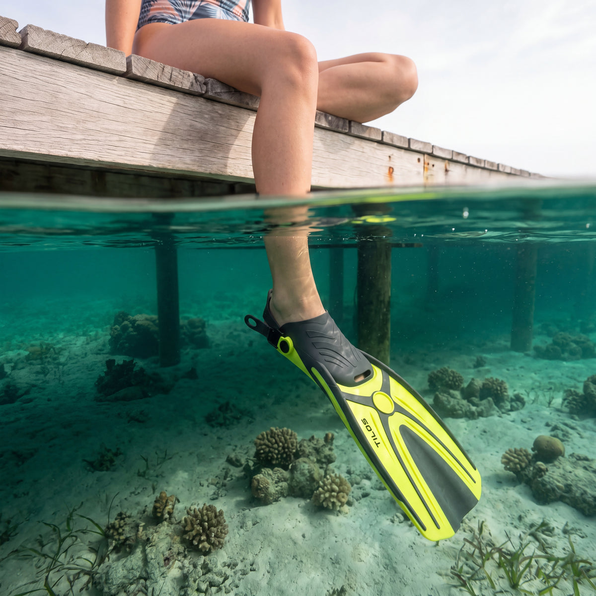 Snorkeler sitting on a wooden dock with one leg hanging into the water, wearing a yellow Tilos Aubade open heel fin