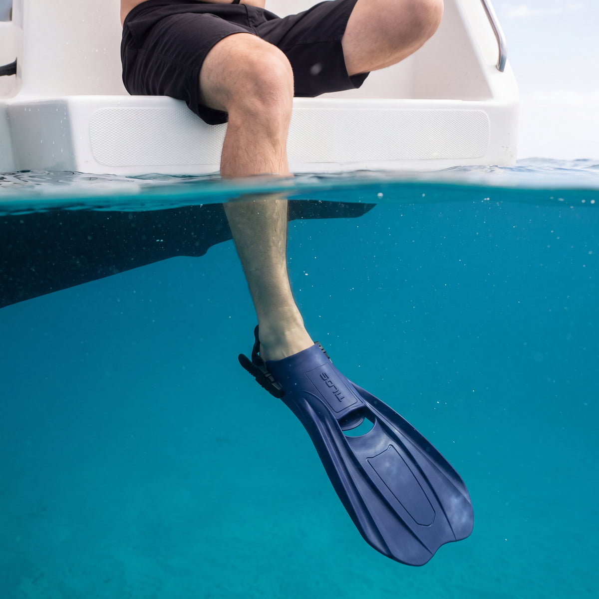 Person sitting on a boat swim step with one leg in clear blue water, wearing a galaxy blue Tilos Technix open heel travel fin