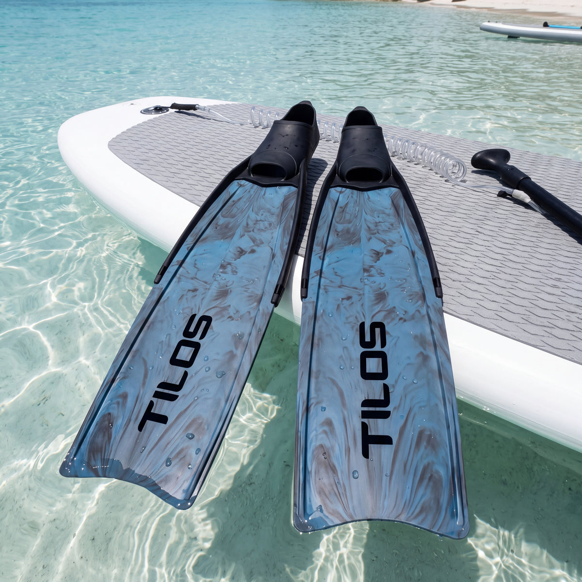 Lifestyle photo of a pair of Tilos Syryn full foot freediving fins in Blue Camo lying on a paddleboard over clear turquoise water