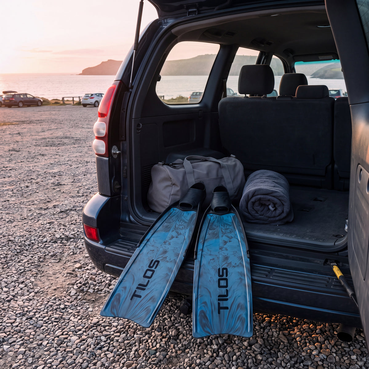 Lifestyle photo of a pair of Tilos Syryn full foot freediving fins in Blue Camo in the back of a car at a coastal car park at sunrise