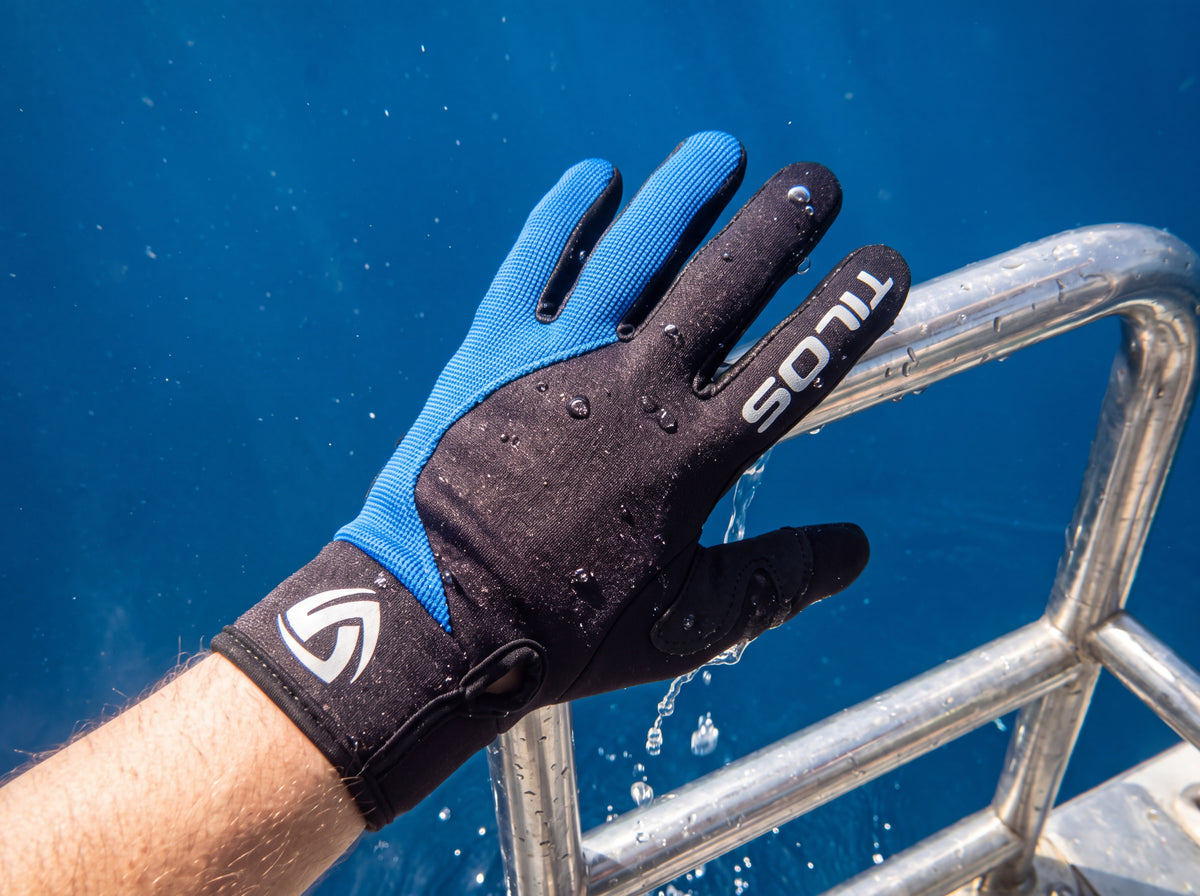 A scuba diver grips the stainless rail of a boat ladder while climbing out of deep blue open water, wearing blue accented Tilos 1.5mm Tropical-X Mesh gloves with seawater streaming off the neoprene.