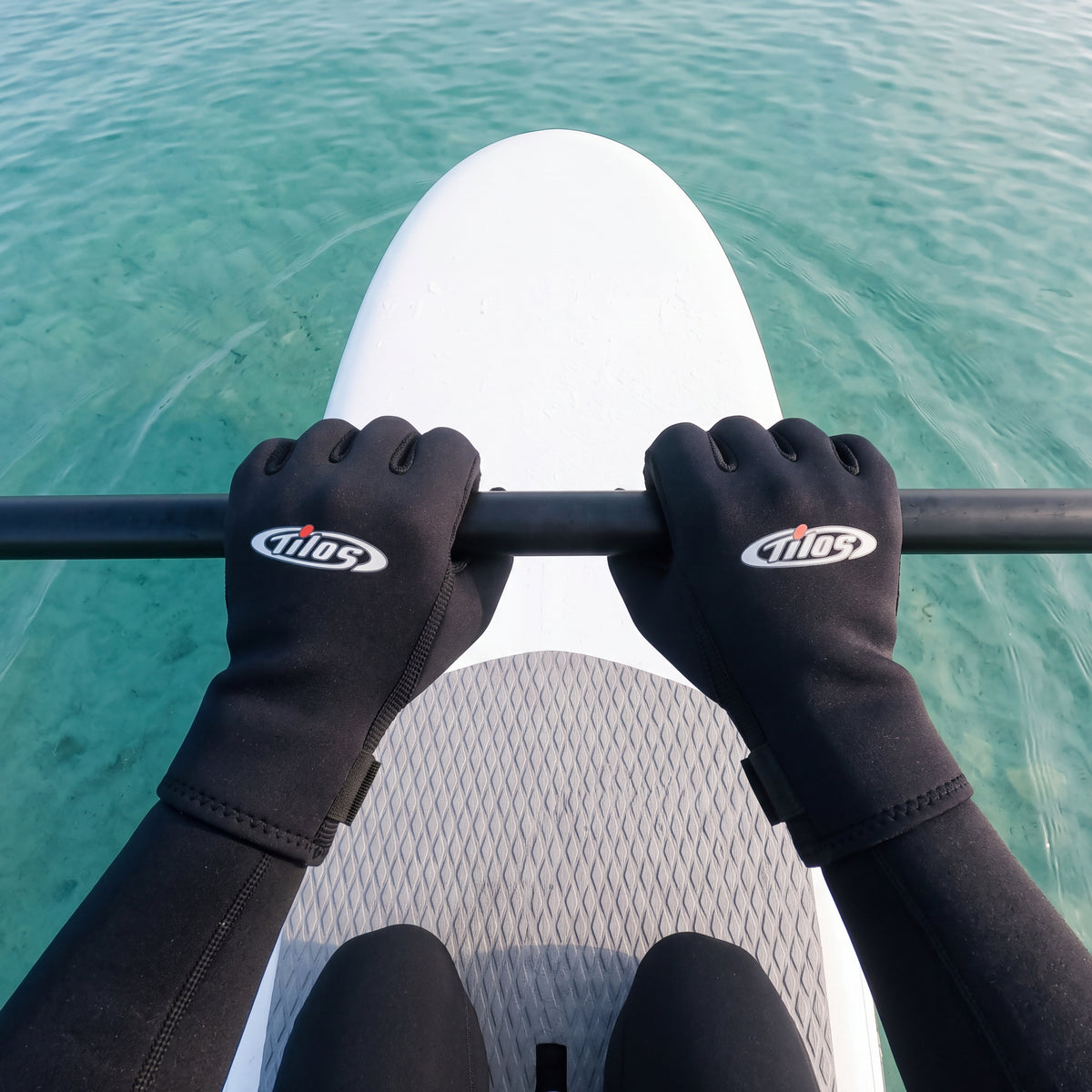 First person view of a paddler kneeling on a paddleboard over clear turquoise water, wearing black Tilos Supratex dive gloves gripping the paddle shaft