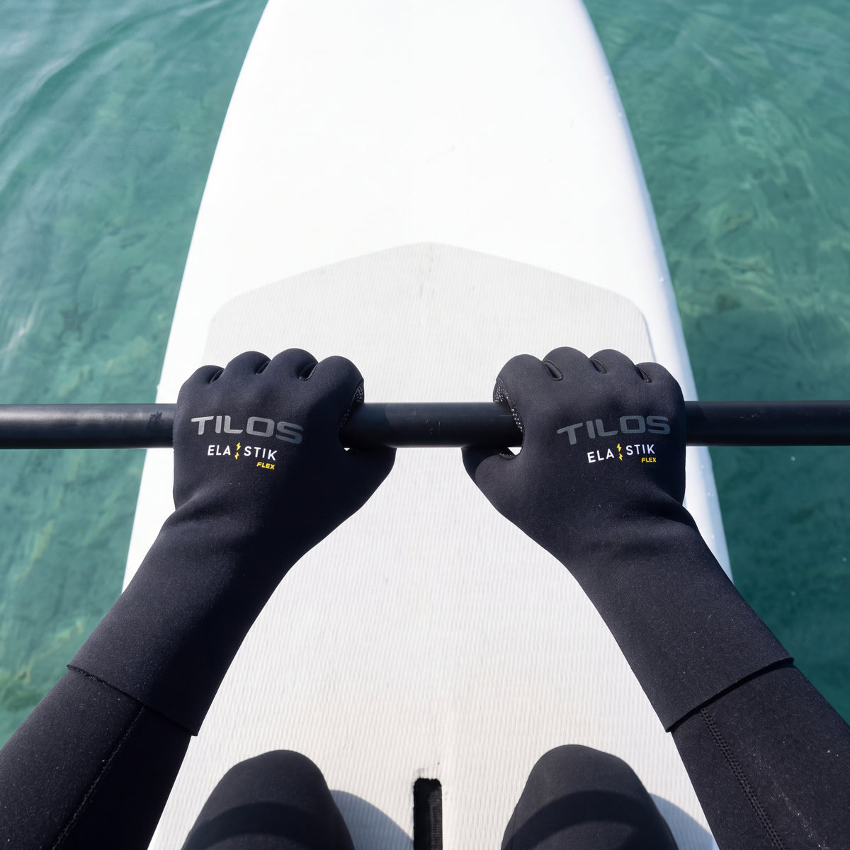 First person view of a paddler kneeling on a white paddleboard over clear turquoise water, wearing black Tilos 2mm Elastik SuperStretch gloves gripping the paddle shaft
