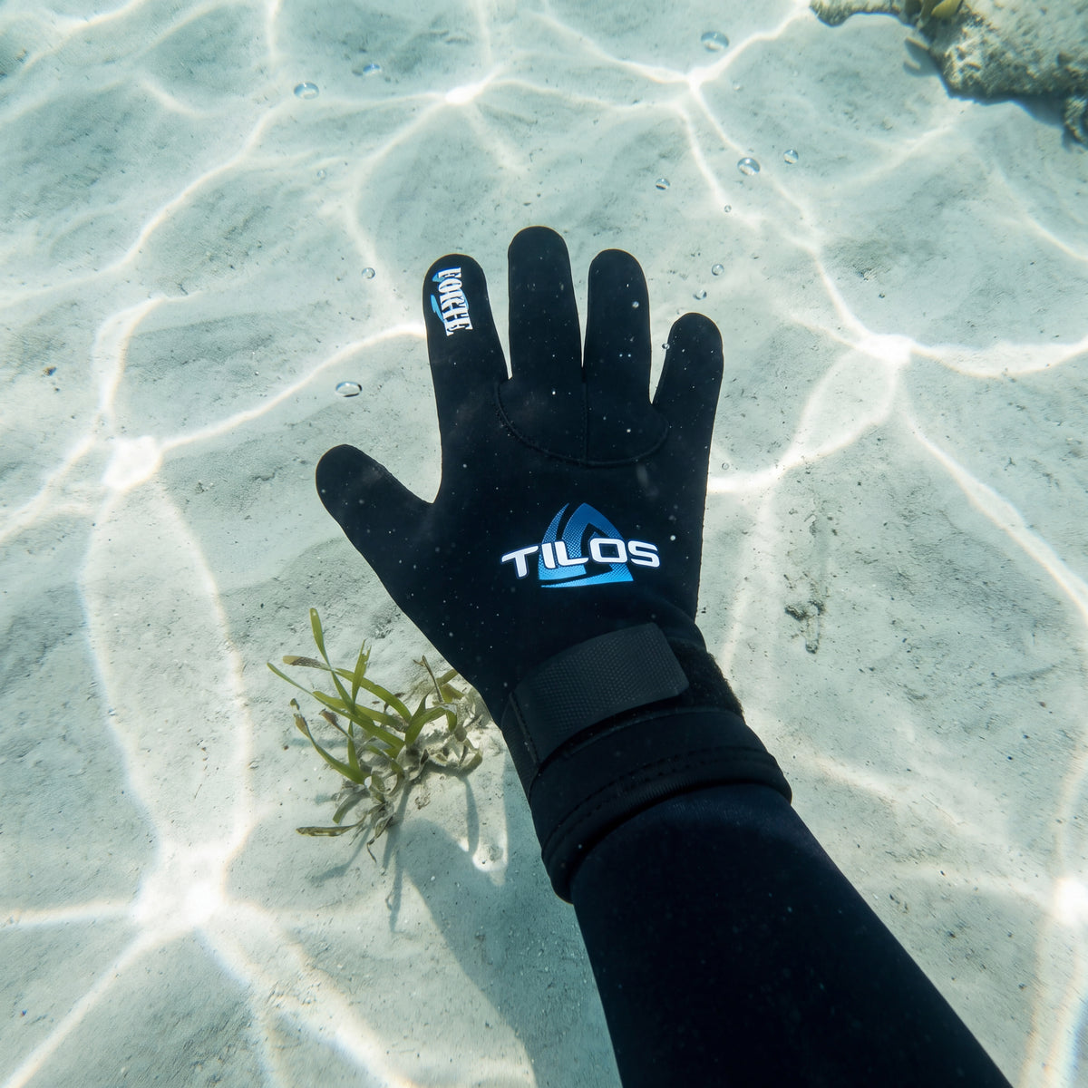 Diver underwater over pale rippled sand wearing a black Tilos 3mm Forte Velcro Glove, hand steadying a hover in shallow sunlit water