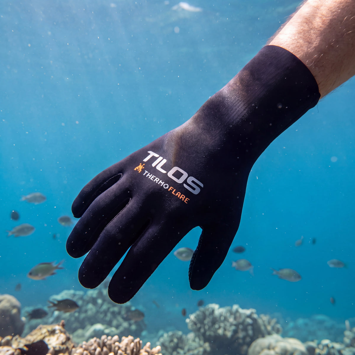 Diver finning through clear blue water with one arm extended forward, wearing black Tilos 3mm Thermoflare superstretch dive gloves underwater.