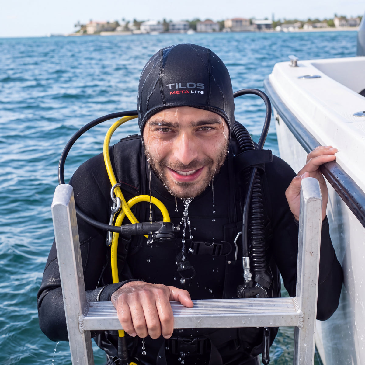 Diver climbing the boat ladder after a dive with seawater running off the cap, wearing a black Tilos 1mm titanium neoprene beanie.