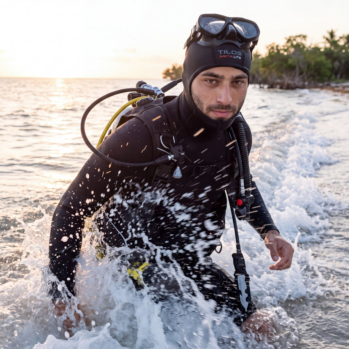 Diver wading out through breaking surf at sunrise on a shore entry, wearing a black Tilos 1mm titanium neoprene beanie.
