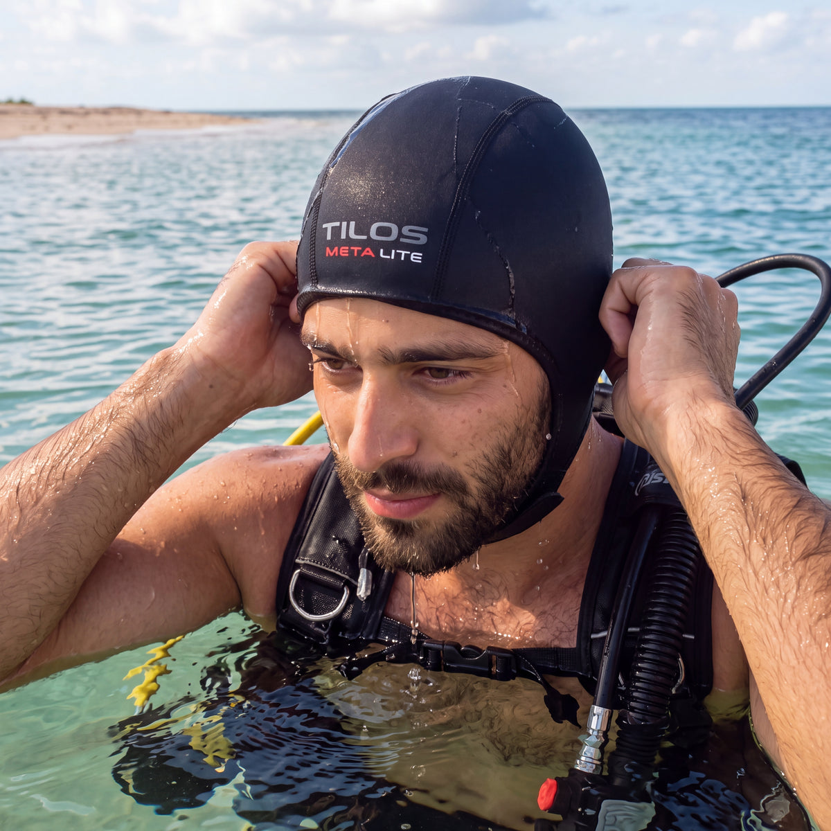 Diver in shallow water pulling the cap down over the ears before a shore dive, wearing a black Tilos 1mm titanium neoprene beanie.