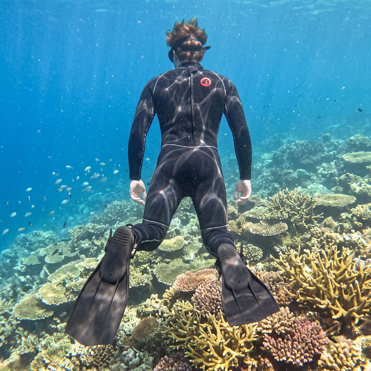 Diver finning away underwater over a shallow coral reef wearing a black Tilos J3204BK full wetsuit and fins.