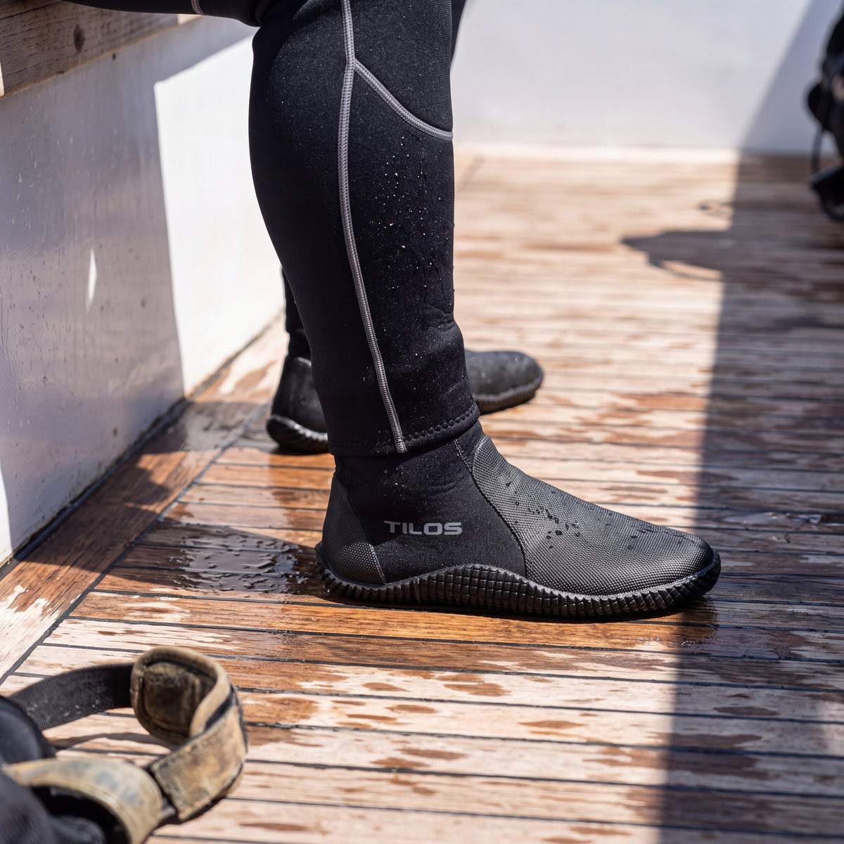 Lifestyle close detail of the integrated neoprene bootie and grey seam piping on a black Tilos J3204BK full wetsuit, on a wet boat deck.