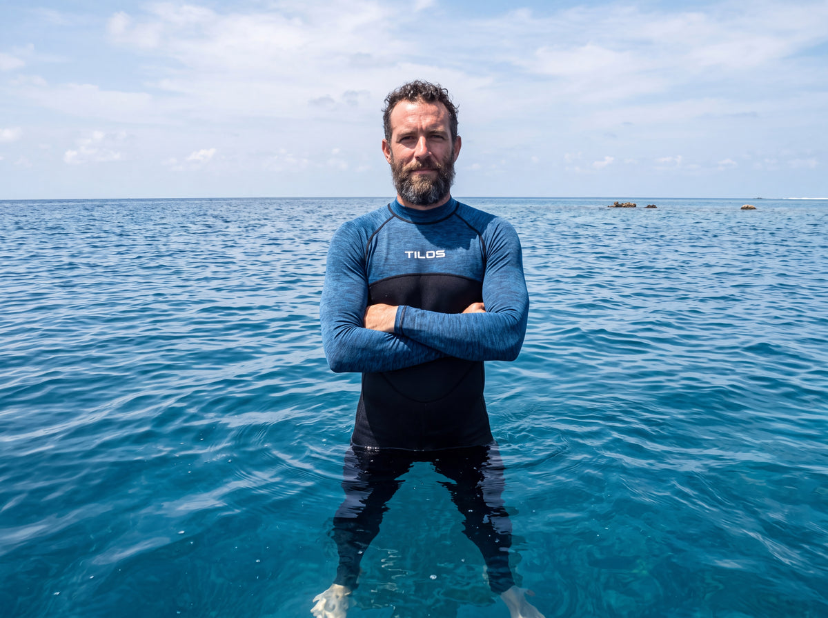 A snorkeler in the Tilos 6oz unisex skin suit in Heather Marine stands chest deep in clear open water with the horizon behind.