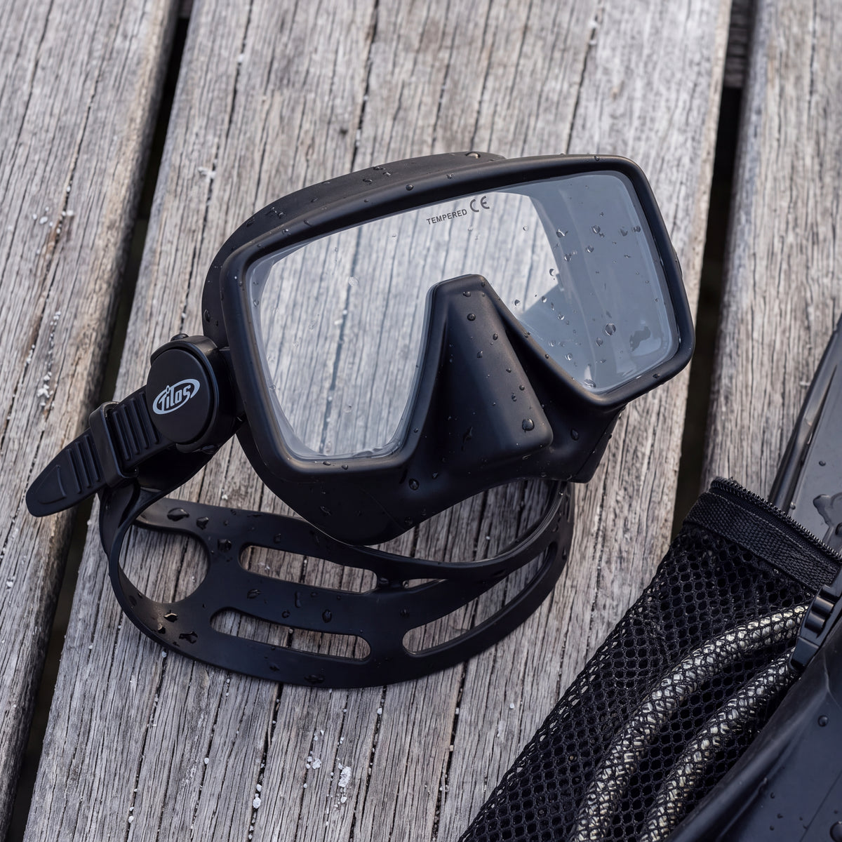 Lifestyle flat lay of the black Tilos Insight Frameless Mask on dock boards beside an open mesh dive bag