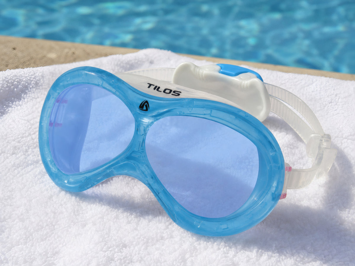 Transparent Blue Tilos Dyno toddler swim mask resting on a folded white towel poolside beside sunlit blue pool water