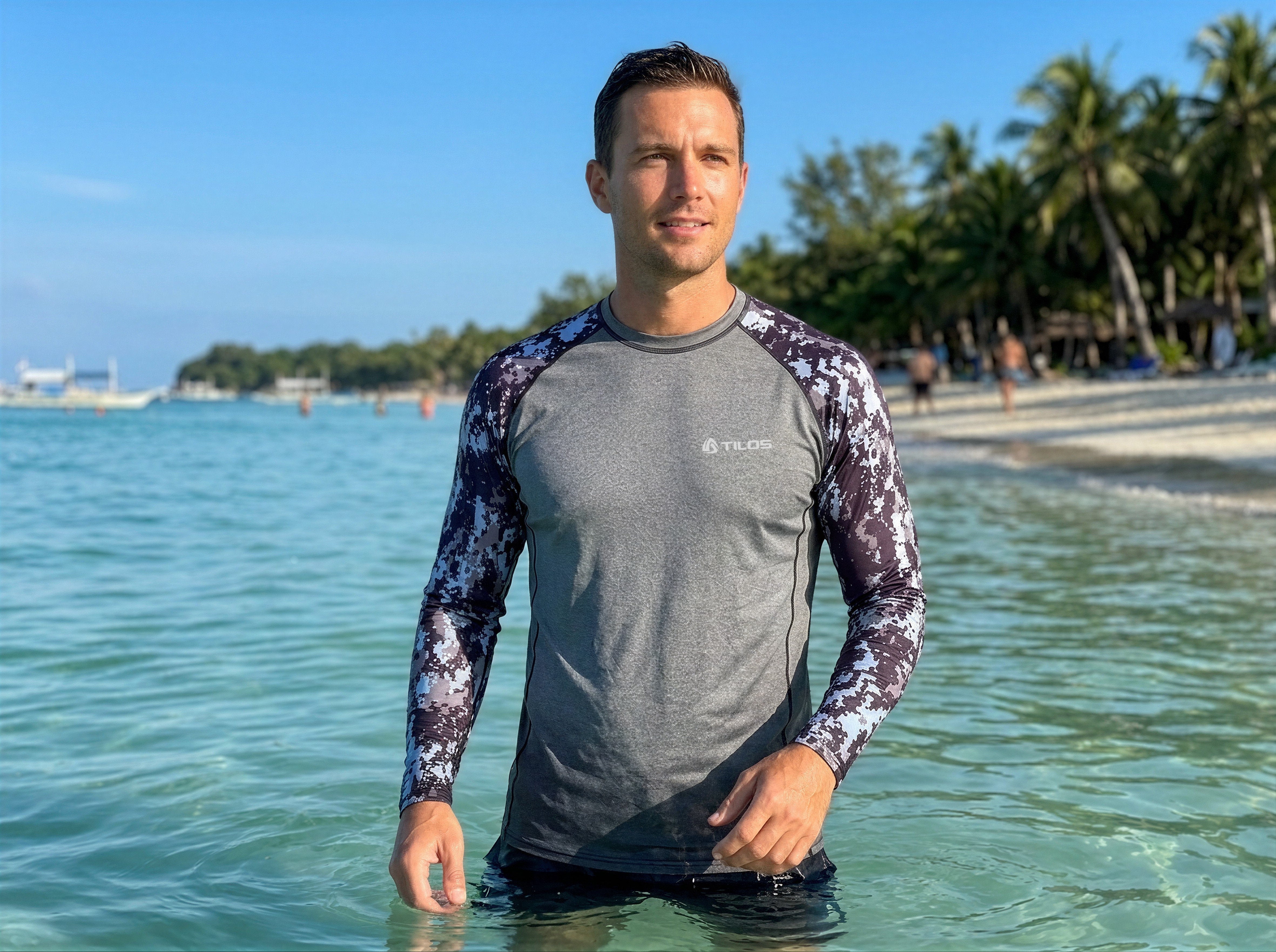Man standing in the ocean at a tropical beach wearing the Tilos Men's 6oz Anti-UV Long Sleeve Rash Guard in Digital Gray camo