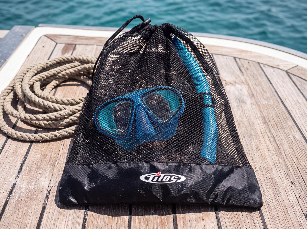 A snorkeler has set a black Tilos mesh mask and snorkel bag on a teak dive boat deck beside a coiled rope.