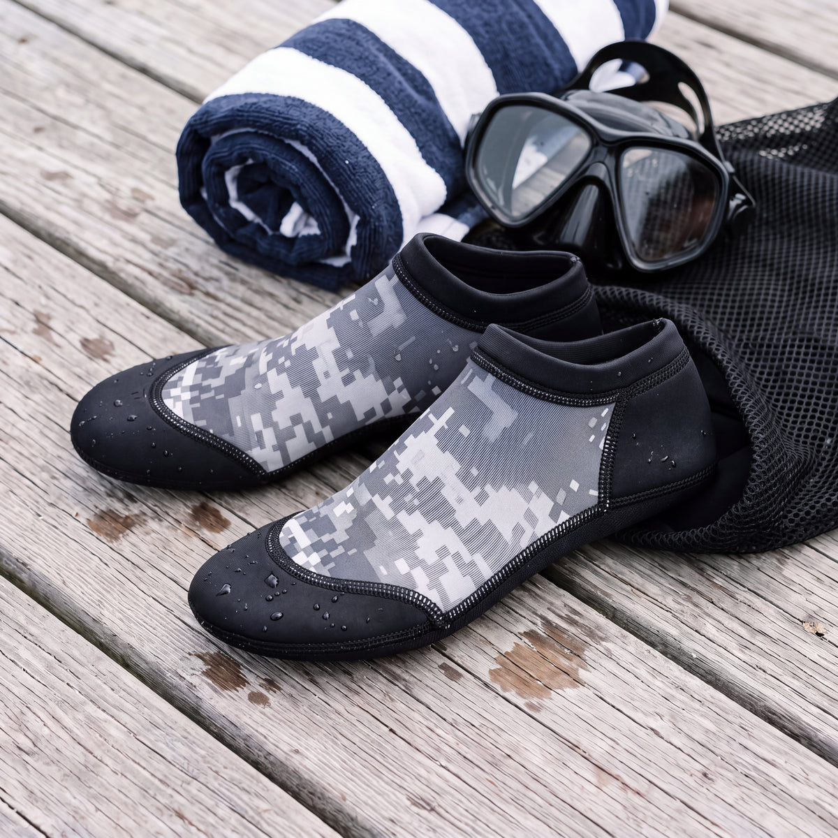 Lifestyle flat lay of the digital gray camo Tilos 2.5mm Low Cut Sport Skin Socks on dock boards beside a dive mask, towel and mesh bag