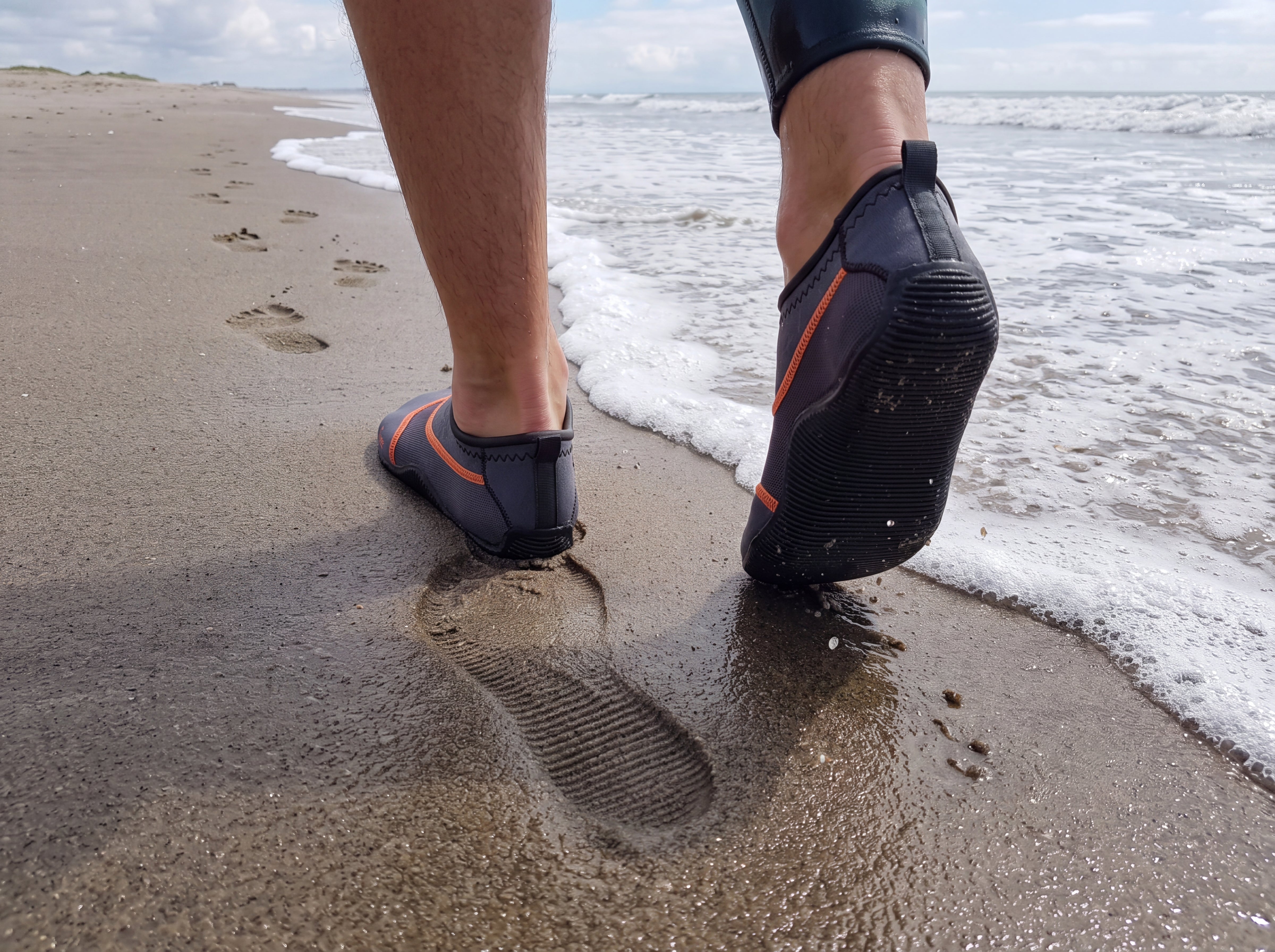Gray Tilos Osmos non-slip water socks being worn walking along a wet sand shoreline, rear view with a ribbed sole imprint left in the sand