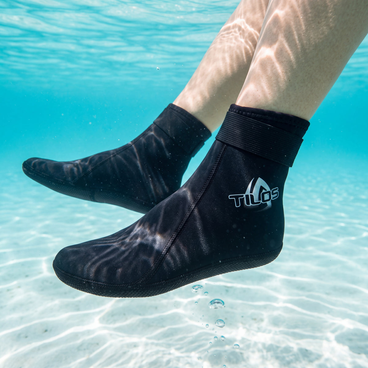 Underwater view of a person floating in a clear turquoise lagoon wearing the black Tilos 2mm Mid-High Top Socks with Velcro Straps