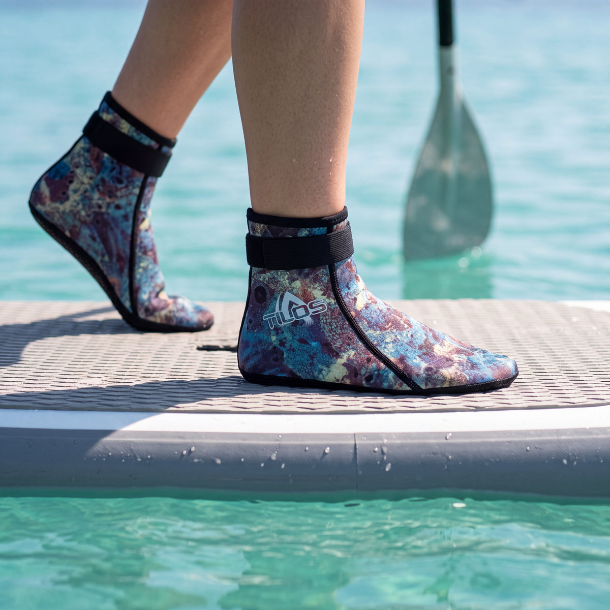 Paddleboarder standing on the deck pad of a stand up paddleboard wearing the green camo Tilos 2mm Mid-High Top Socks with Velcro Straps