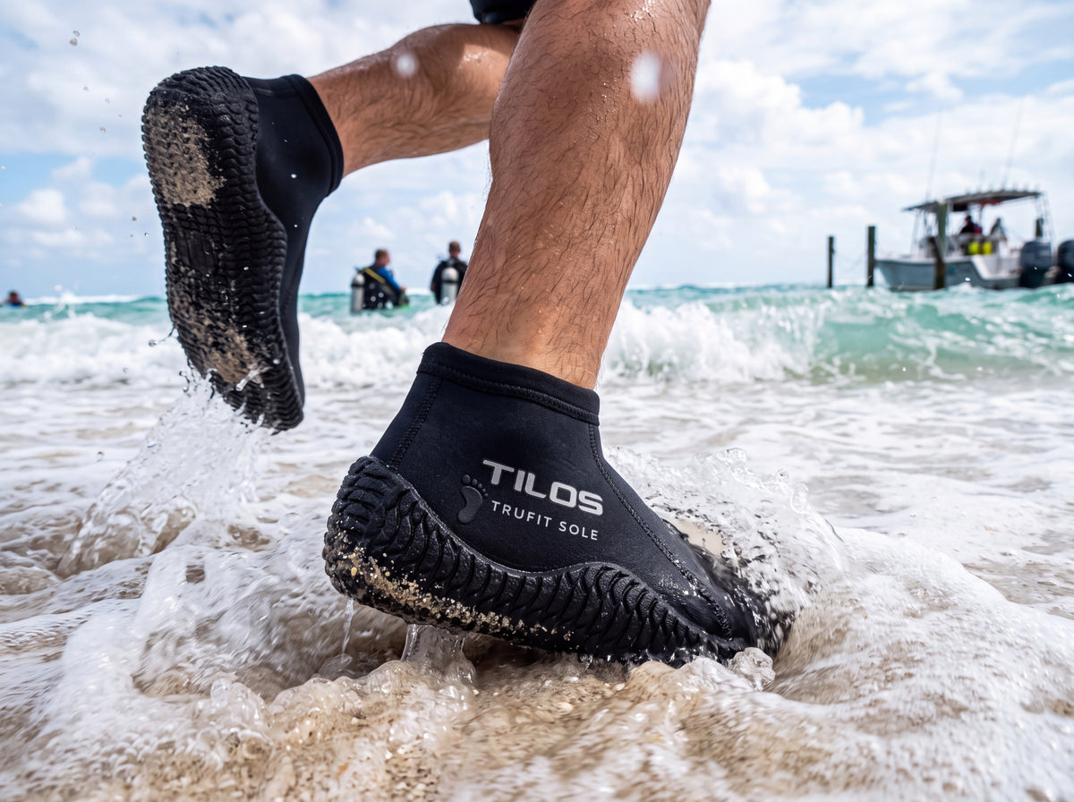 A diver wearing black Tilos 3mm low cut TruFit beach boots runs into the surf entering the water off a sandy beach.