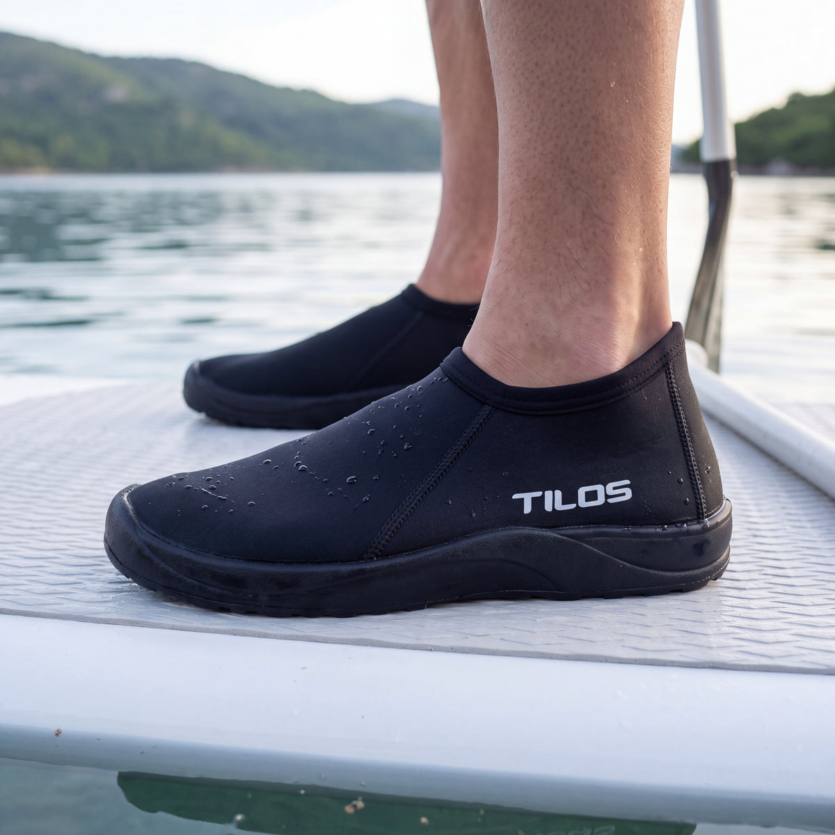 Paddleboarder standing on a white board in a calm bay while wearing the black Tilos 3mm Low Cut Molded Vulcanized Sole Beach Boot