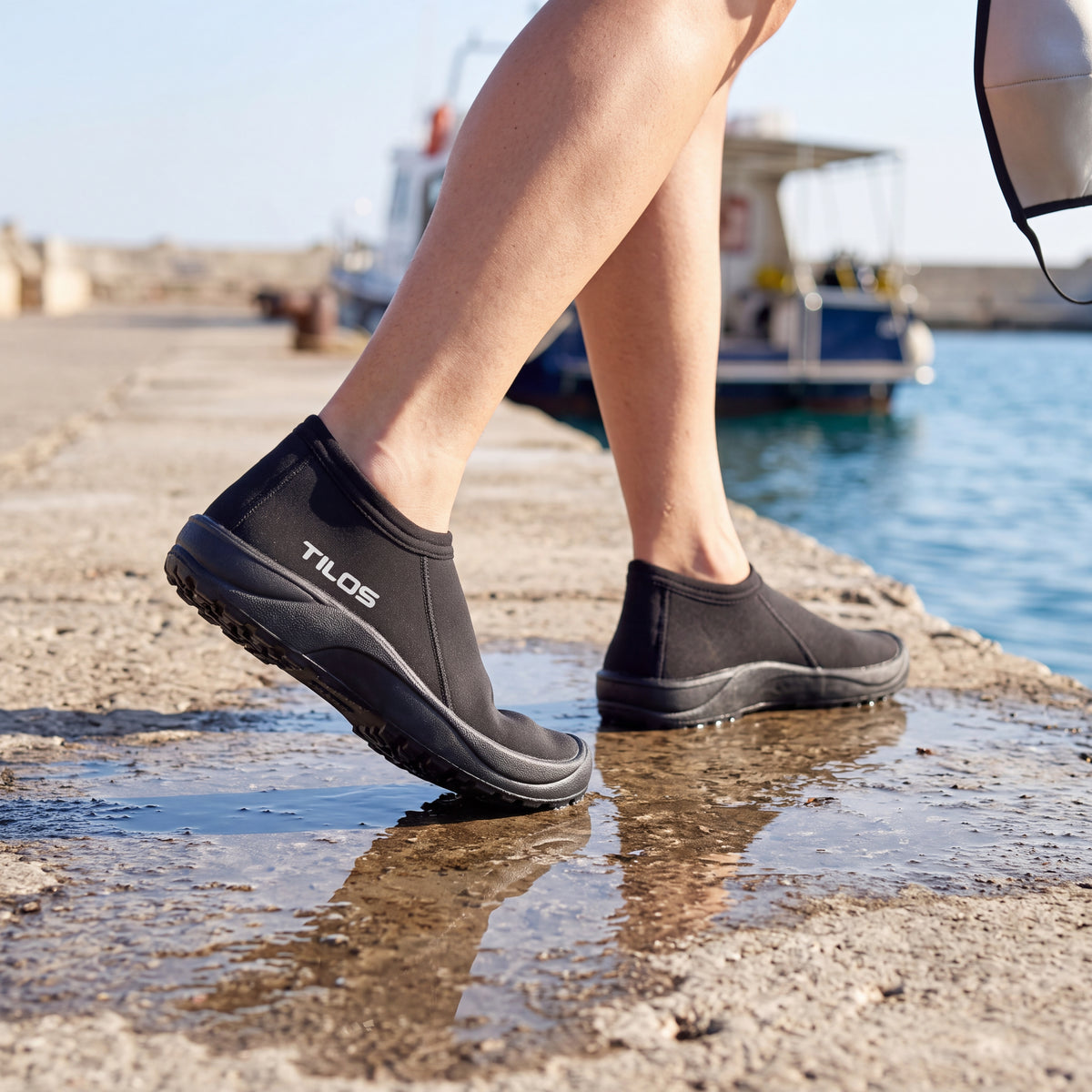 Diver walking along a wet harbour jetty toward a dive boat while wearing the black Tilos 3mm Low Cut Molded Vulcanized Sole Beach Boot