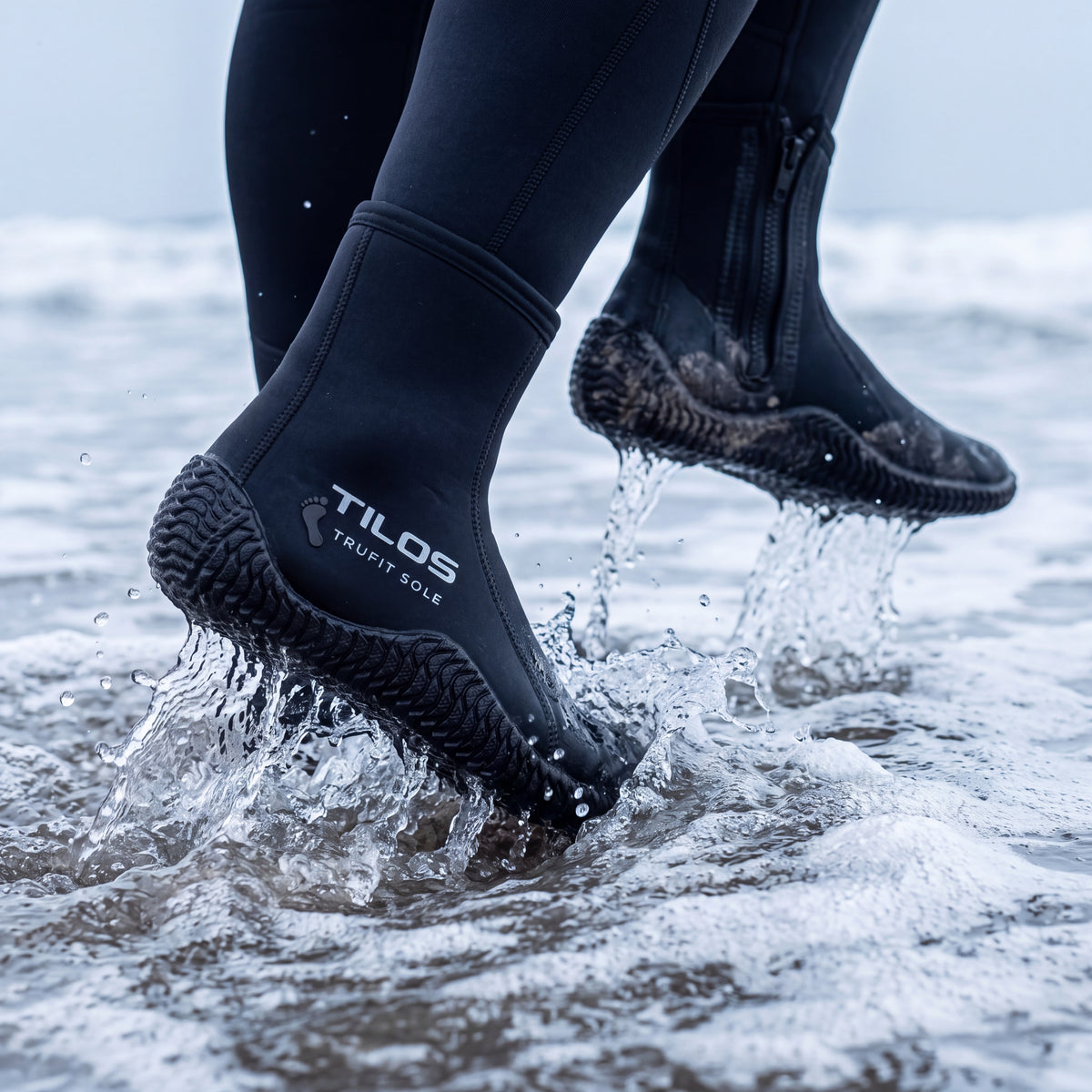 A diver wearing Tilos 5mm Titanium Zip Trufit boots strides head-on through breaking shore break, one boot driving into the whitewater and throwing spray.