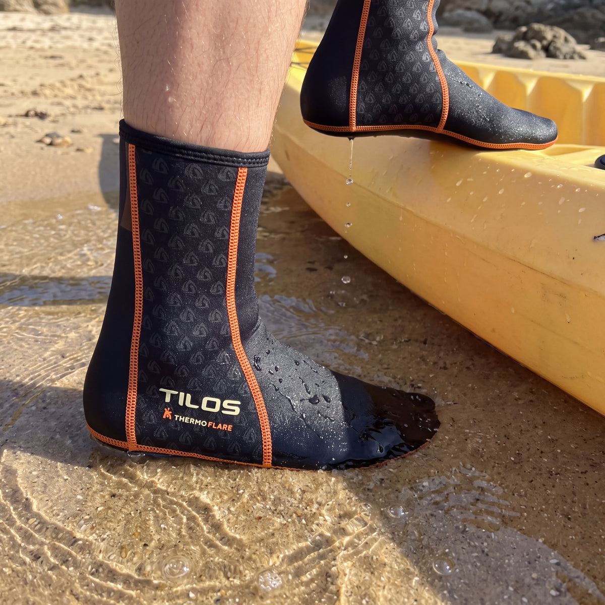Person stepping out of a yellow kayak into shallow water at a sandy cove while wearing the black Tilos 1mm Thermoflare Socks
