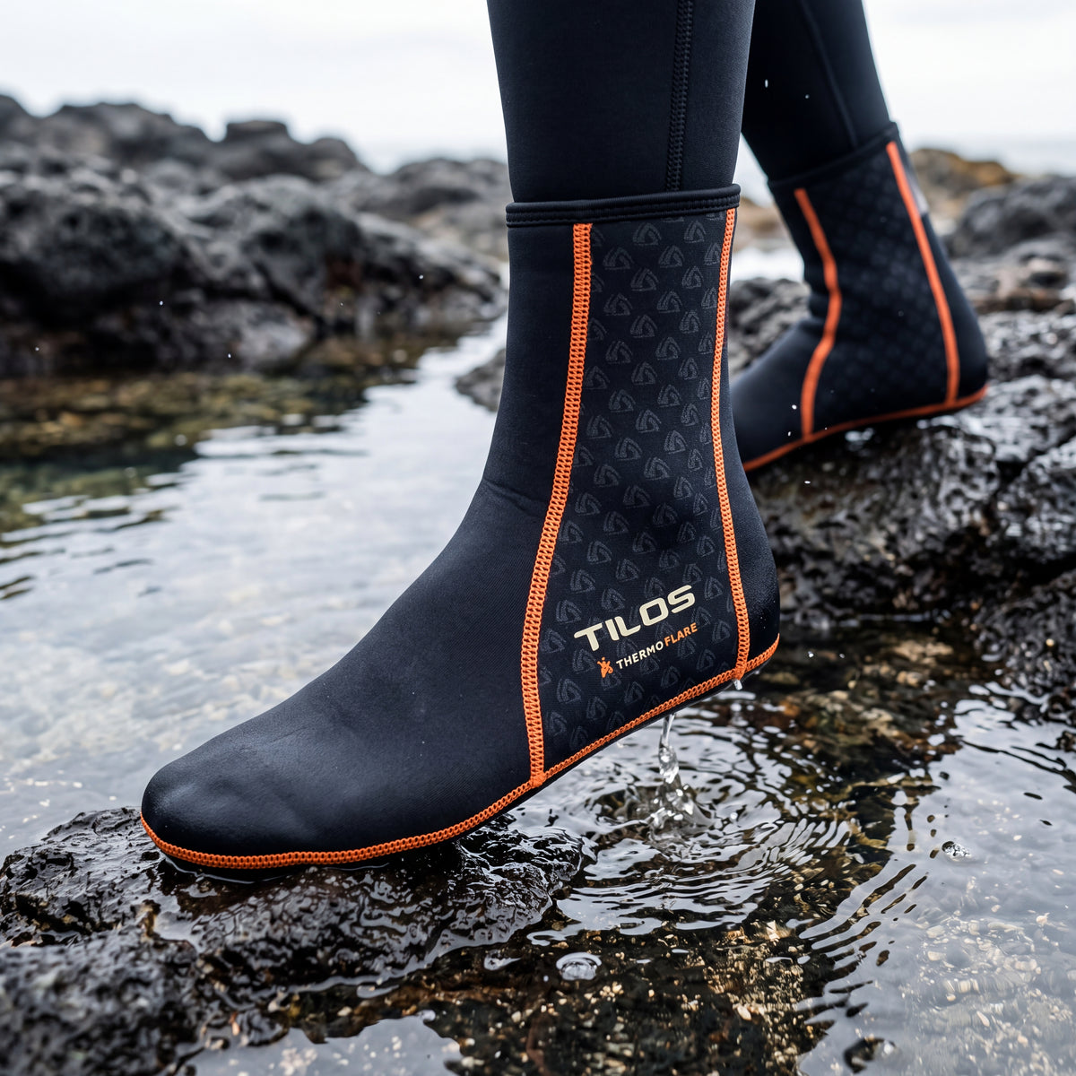 Person stepping across wet volcanic rocks beside a tide pool while wearing the black Tilos 1mm Thermoflare Socks