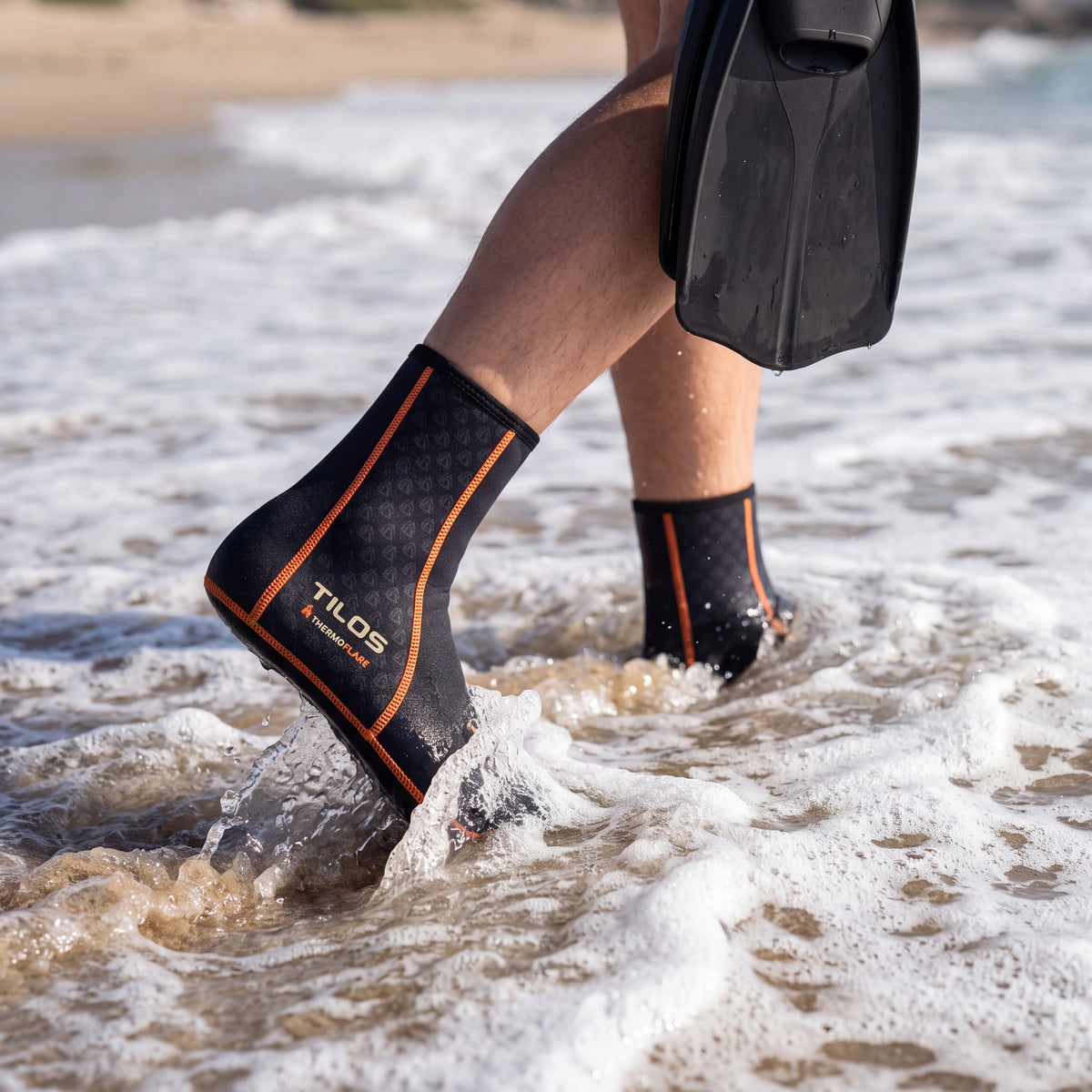 Person wading into the surf carrying fins while wearing the black Tilos 1mm Thermoflare Socks