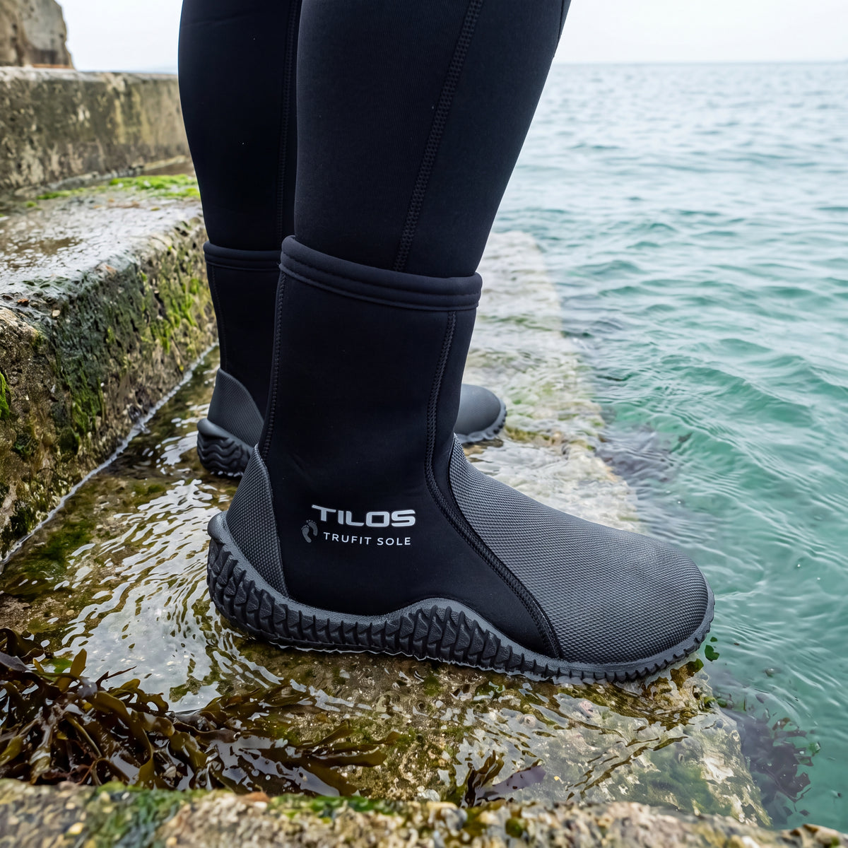 Diver standing on wet stone harbour steps at the water's edge while wearing the black Tilos 5mm Thermoflare Semi-Dry Trufit Boot