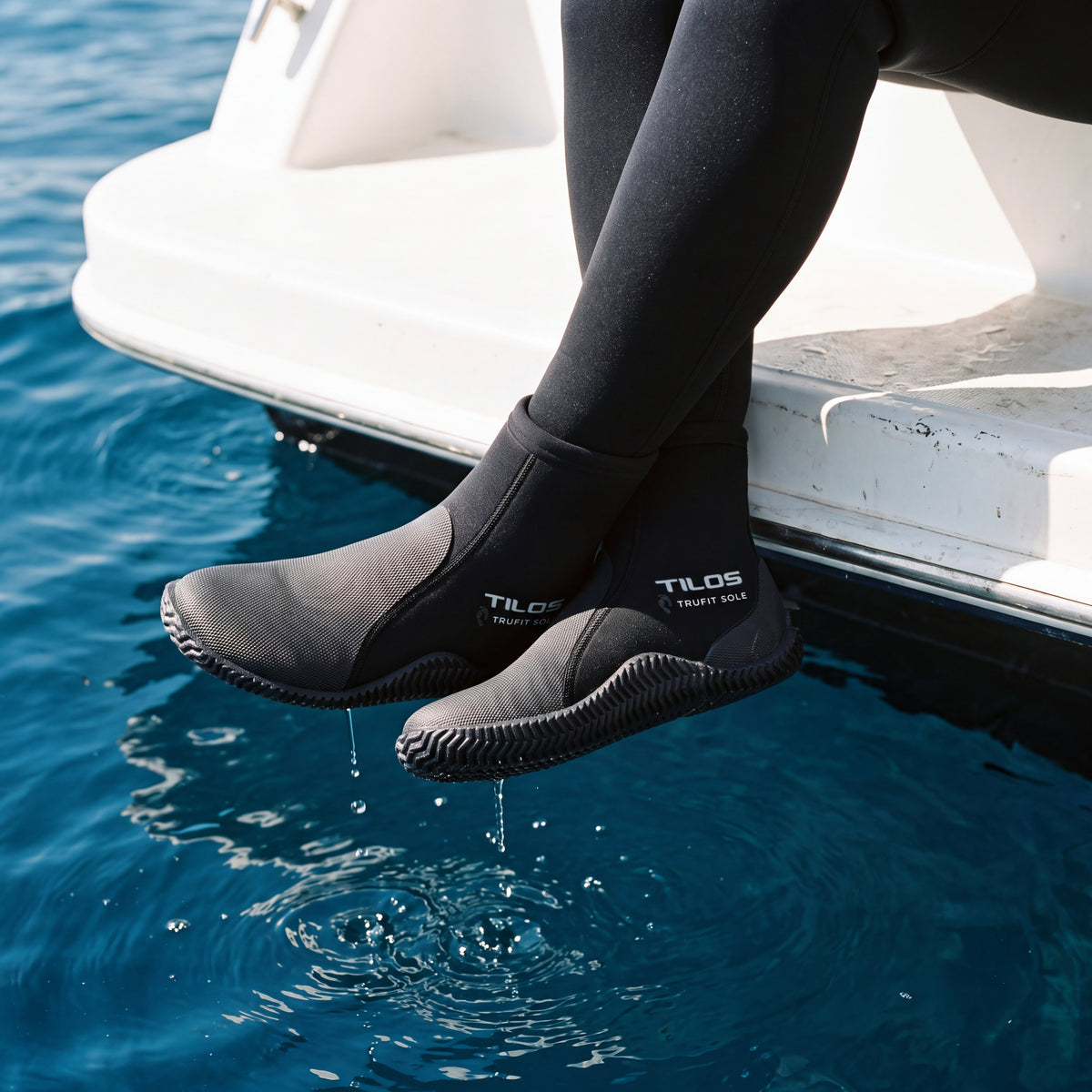 Diver sitting on a boat swim platform above blue water while wearing the black Tilos 5mm Thermoflare Semi-Dry Trufit Boot