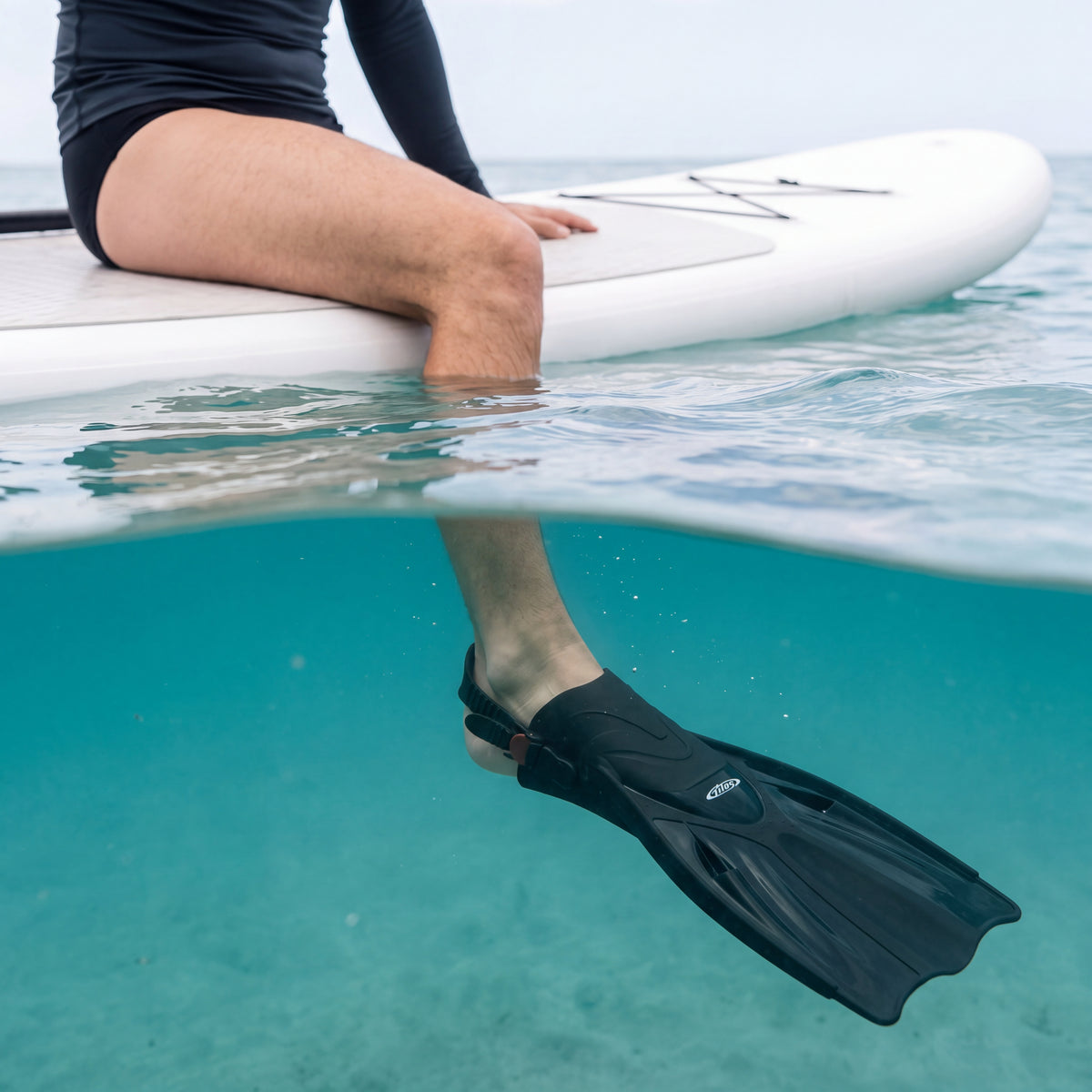 Person sitting on a paddleboard with one leg hanging into clear turquoise water, wearing a black Tilos Getaway open heel fin