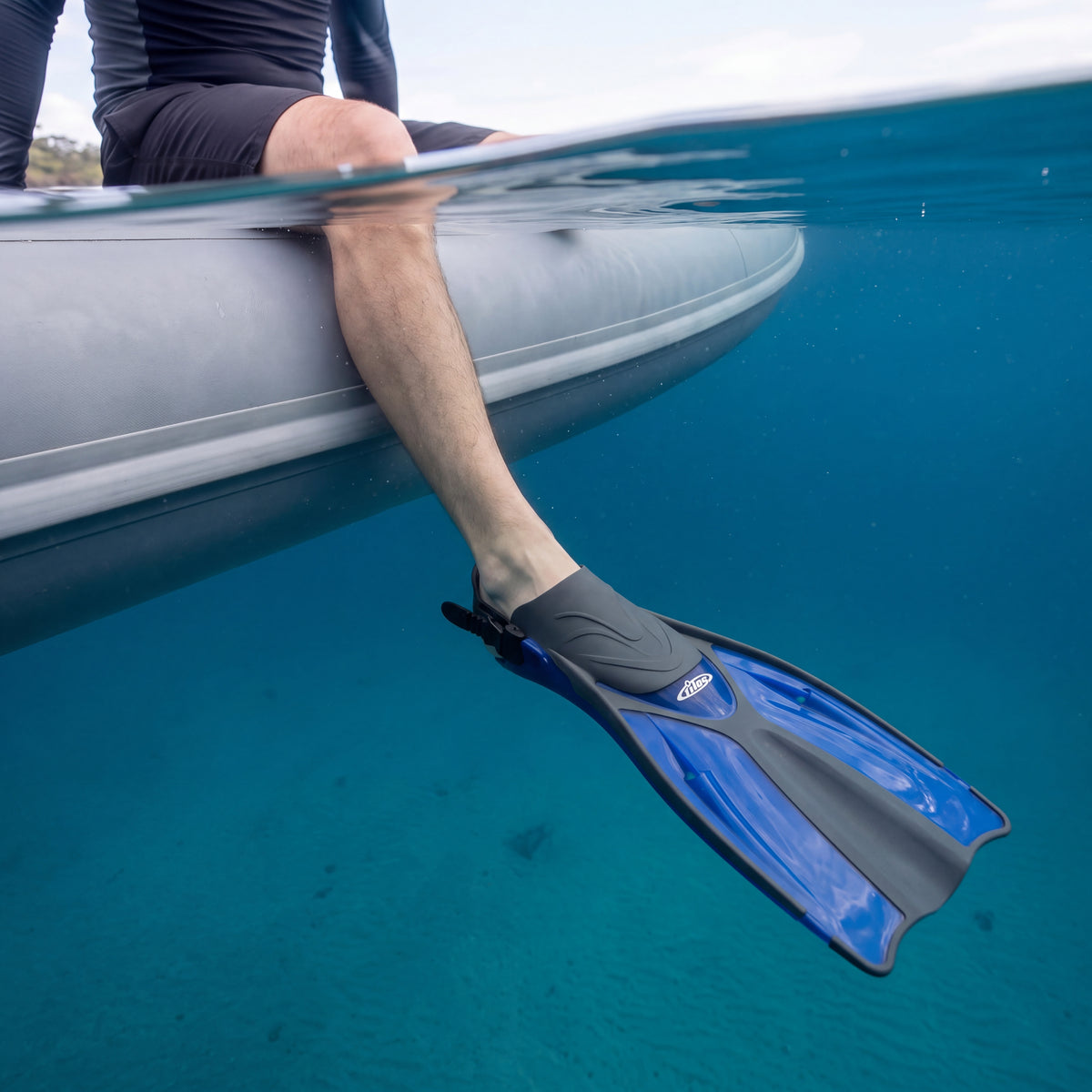 Diver sitting on the side of an inflatable boat with one leg in deep blue water, wearing a blue Tilos Getaway open heel fin