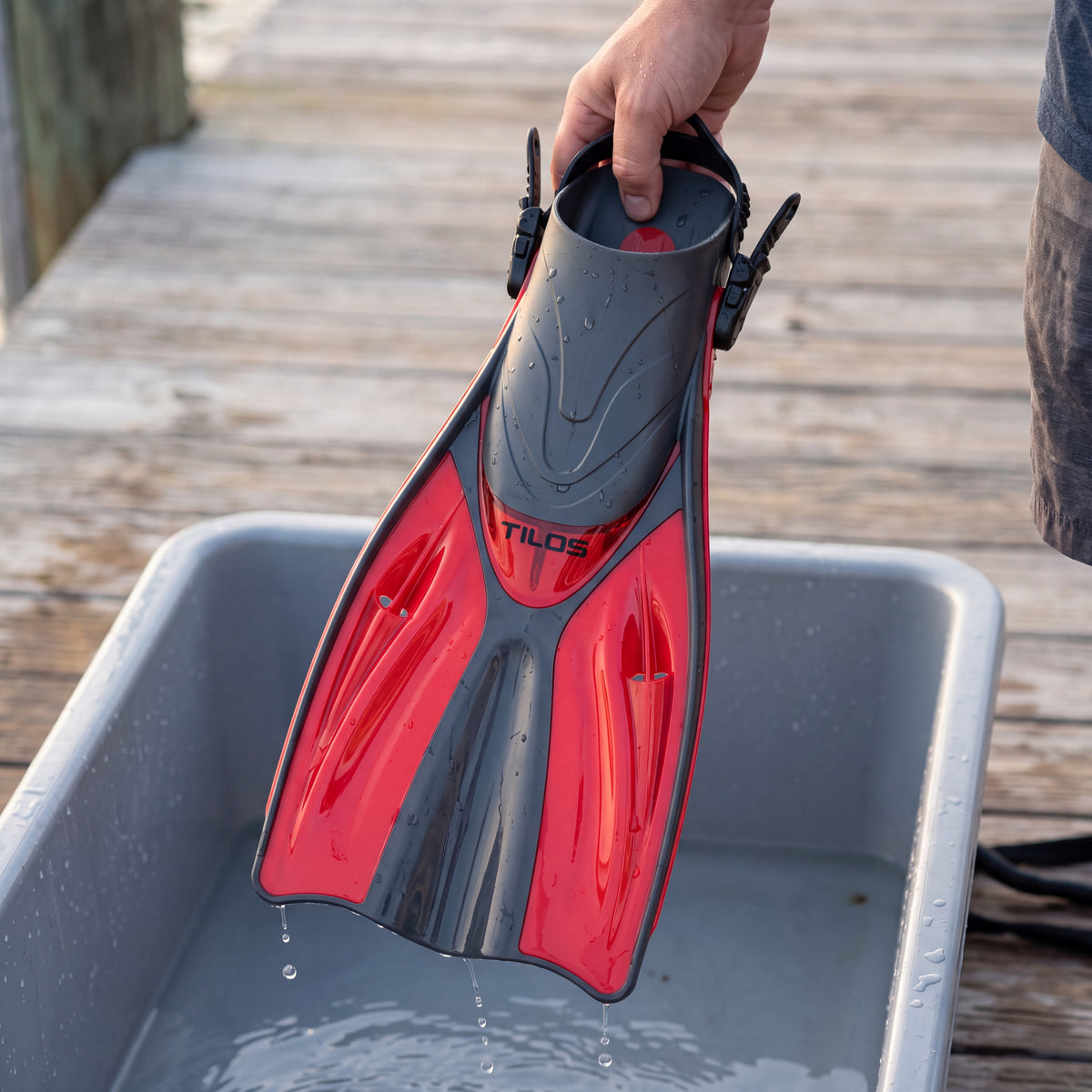 Lifestyle photo of a diver holding a wet red Tilos Getaway open heel fin over a freshwater rinse tub on a dock after a dive