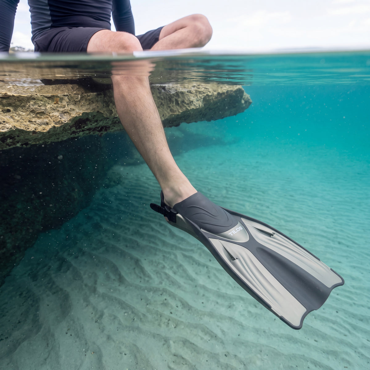 Snorkeler sitting on a rock ledge with one leg in a clear turquoise pool, wearing a titanium Tilos Getaway open heel fin