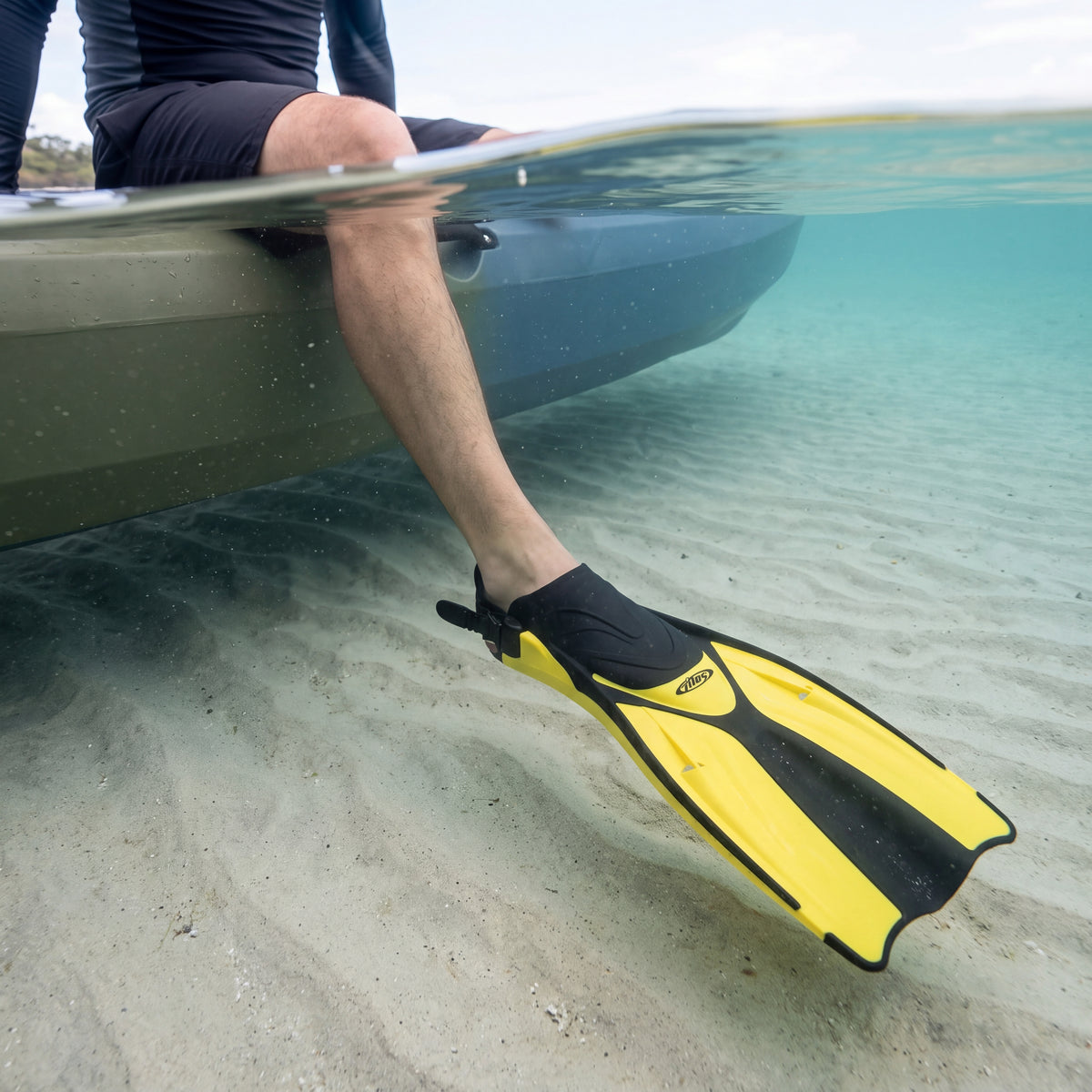 Kayaker sitting on the edge of a kayak with one leg in clear shallow water over sand, wearing a yellow Tilos Getaway open heel fin