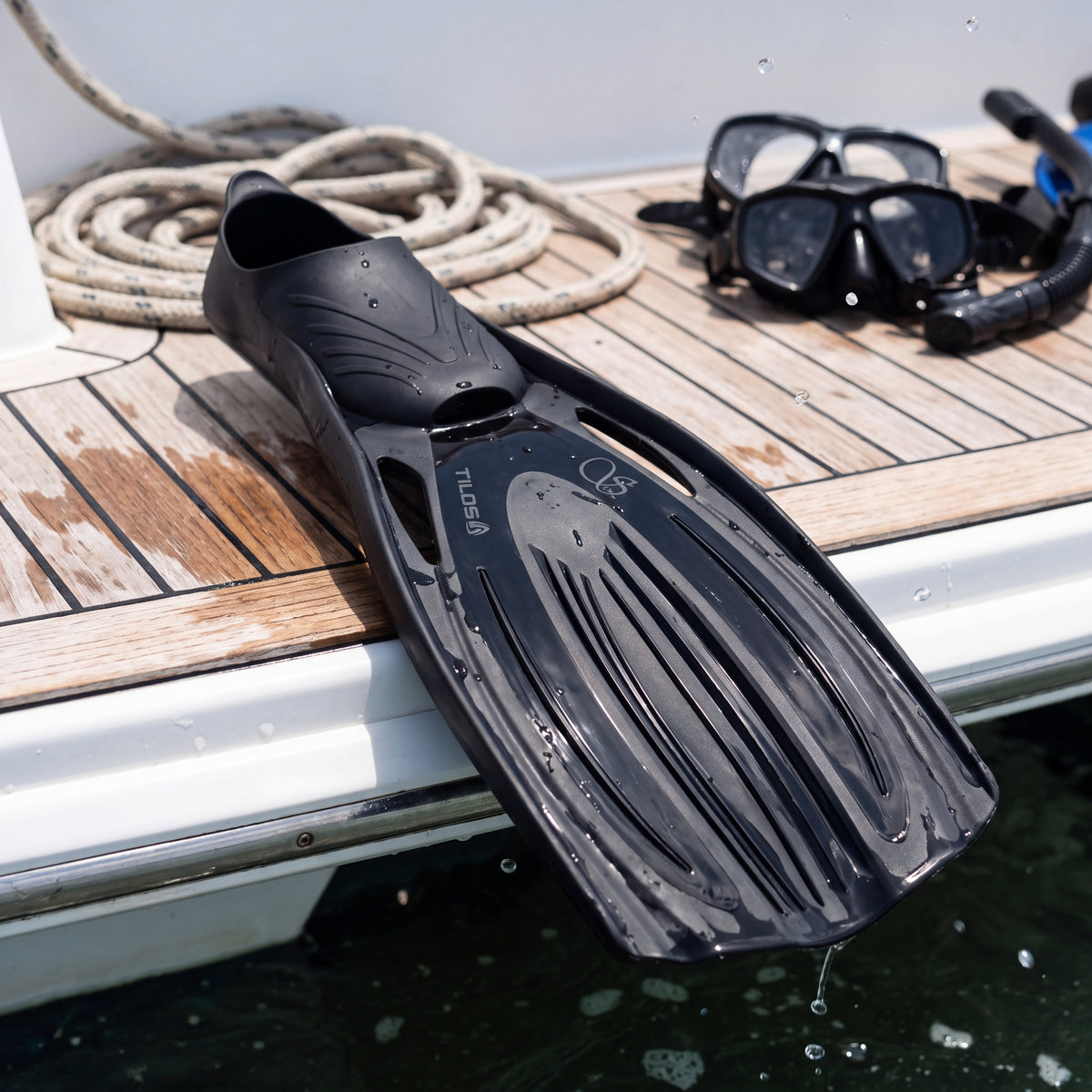 Lifestyle close detail of the Tilos Saber full-foot fin in black resting on a wet teak boat swim step after a dive