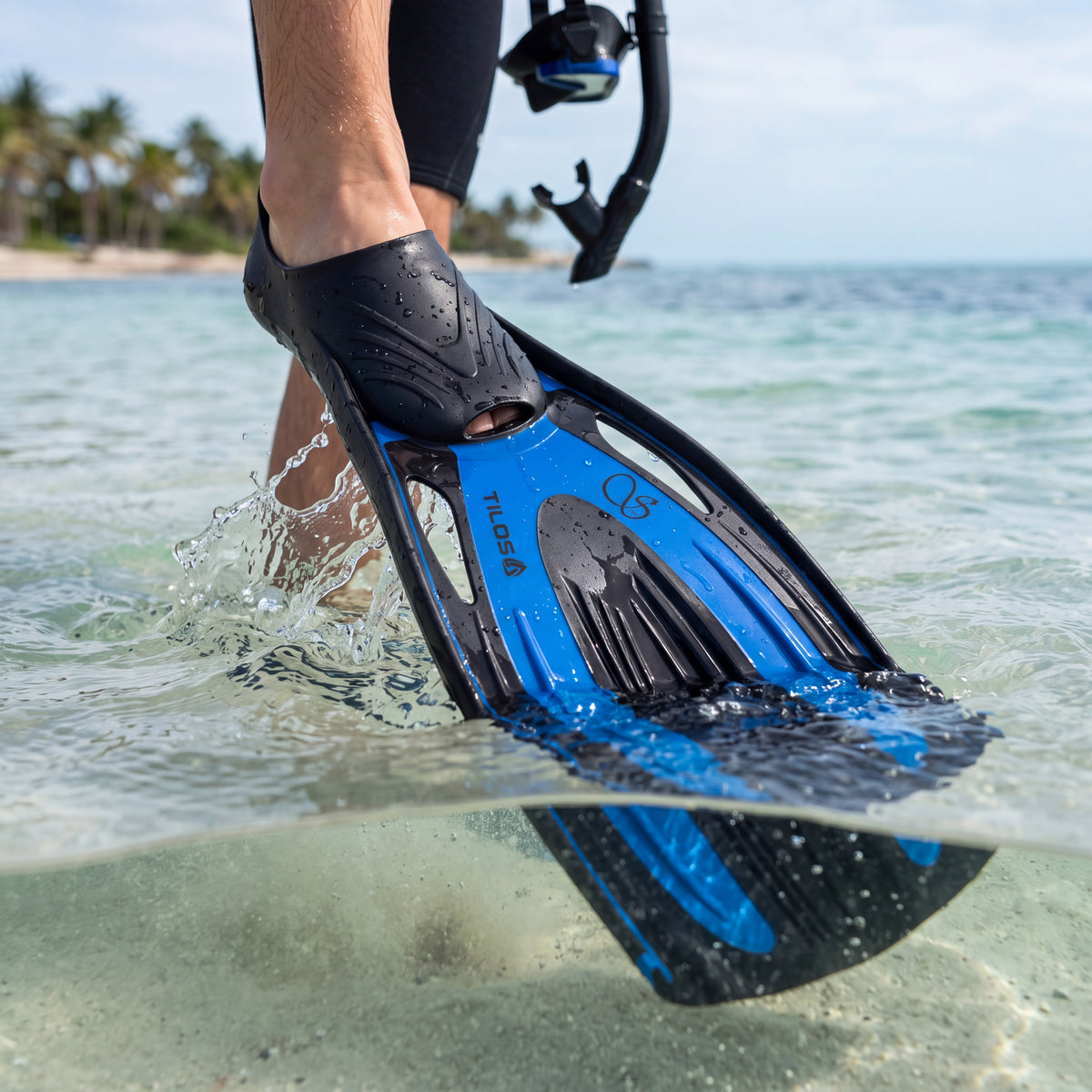 Snorkeler striding out through shallow shore break wearing the Tilos Saber full-foot fins in blue, spray coming off the raised blade