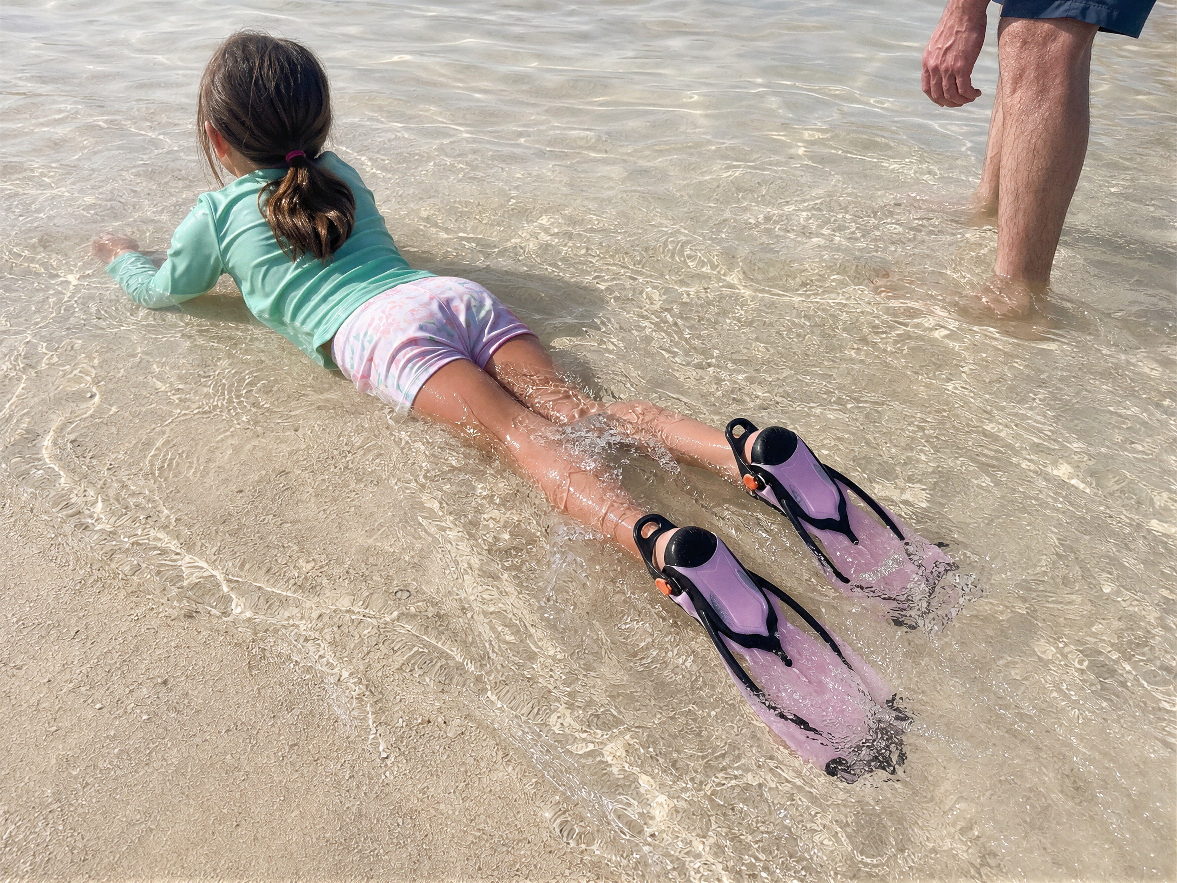 Lifestyle image of a child wearing pink Tilos Vulcan Jr fins in shallow beach water with adult supervision