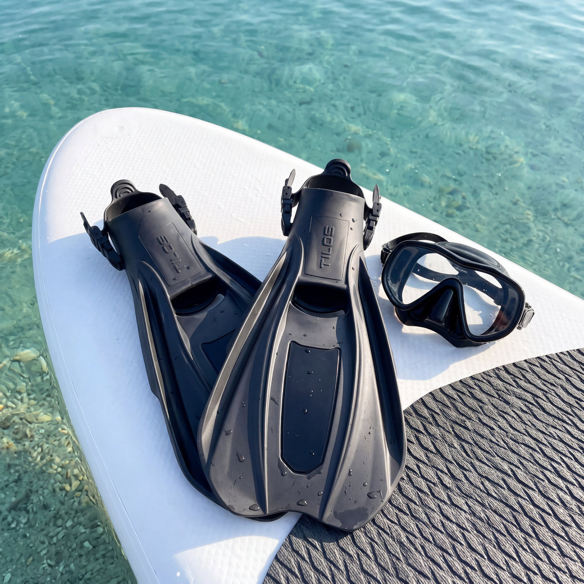 Lifestyle photo of a pair of black Tilos Technix open heel travel fins and a snorkel mask on a paddleboard over clear shallow water